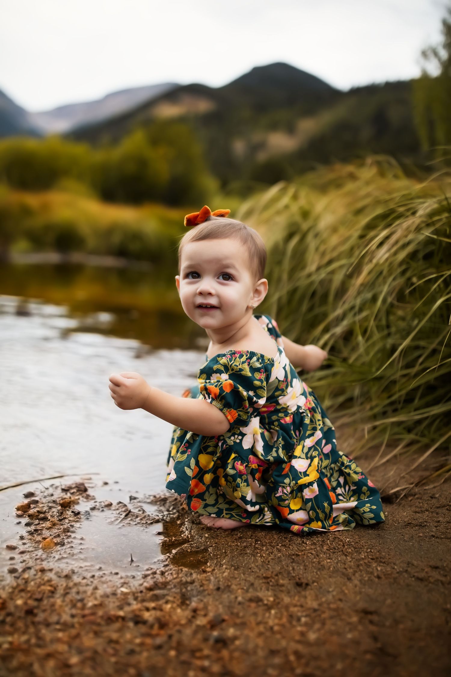 Baby in floral dress plays by riverbank with mountains and autumn foliage in background.