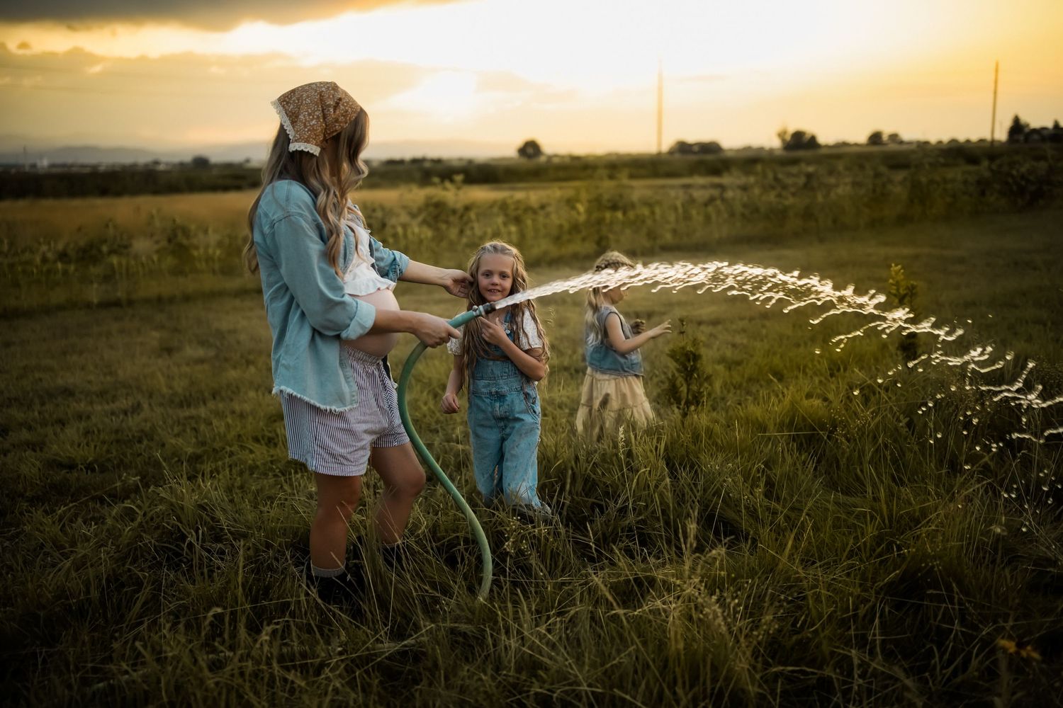 People playing with water from a garden hose in a golden sunset-lit field during summertime.