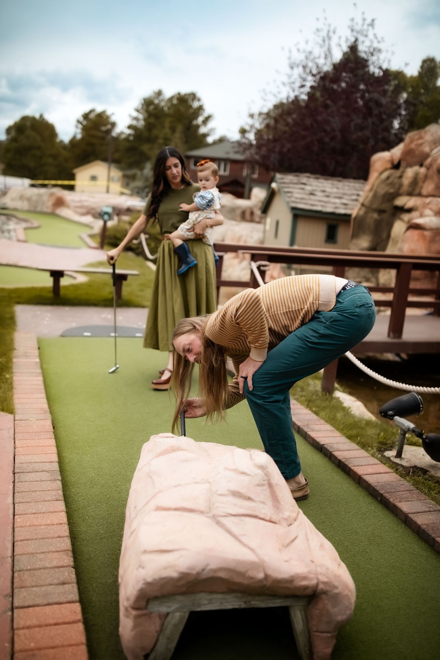 People playing miniature golf on a course with one person leaning over to retrieve something from a bench.
