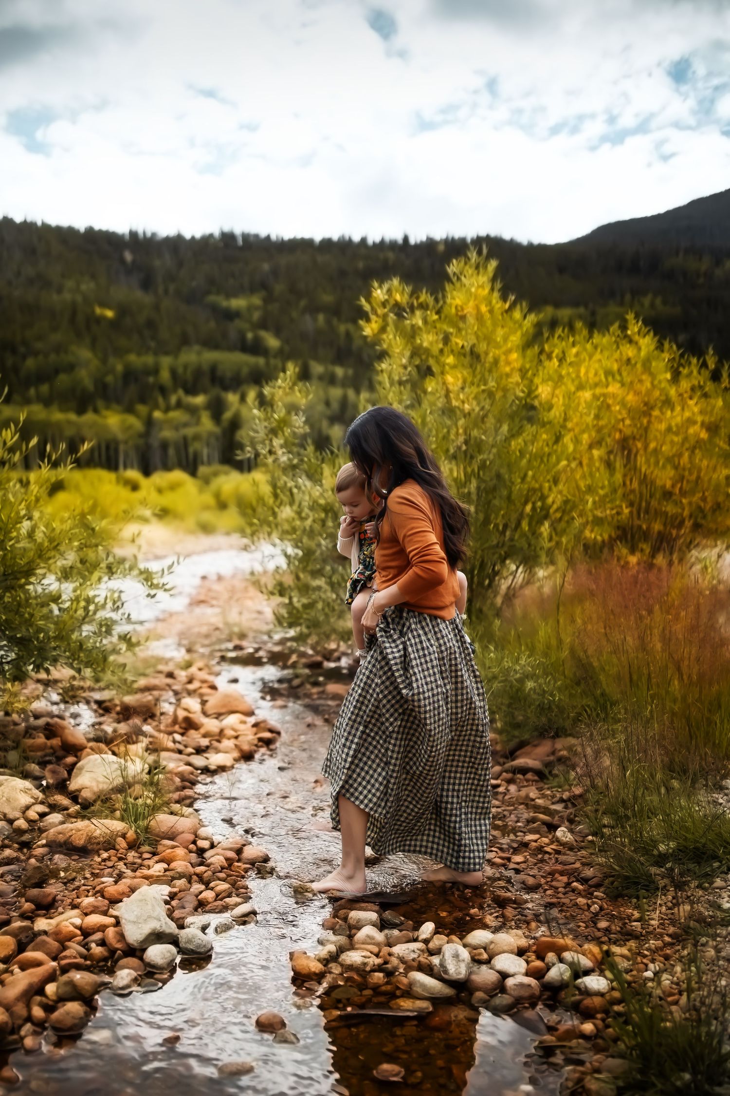 Two people sit among rocks by a stream with mountains and yellow foliage in the background.
