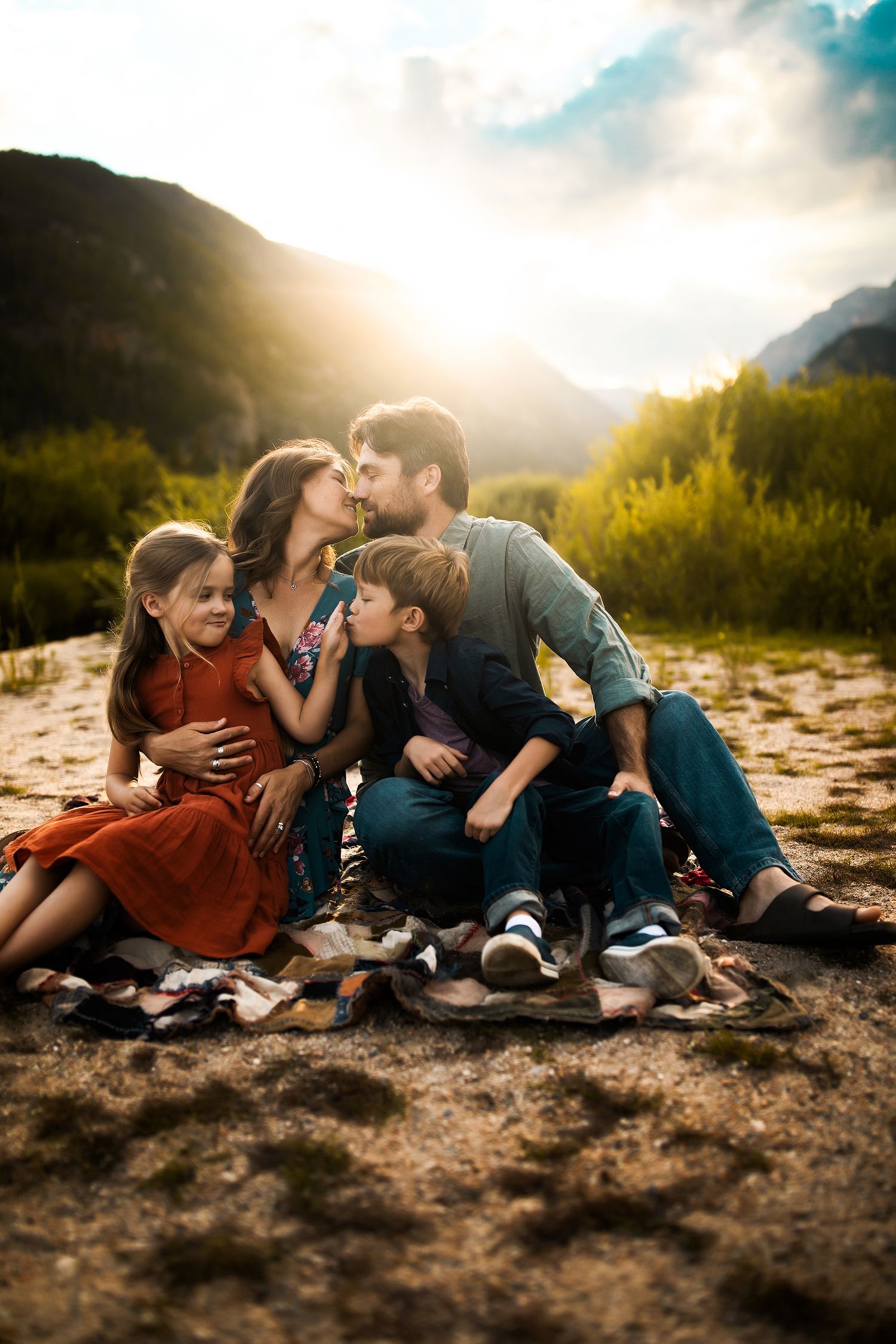 Family of four sitting on a blanket outdoors at sunset, with mountains and greenery in the background.