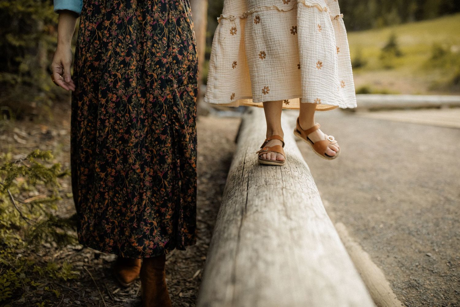 Woman and child walking on wooden beam, wearing floral skirts and sandals outdoors.