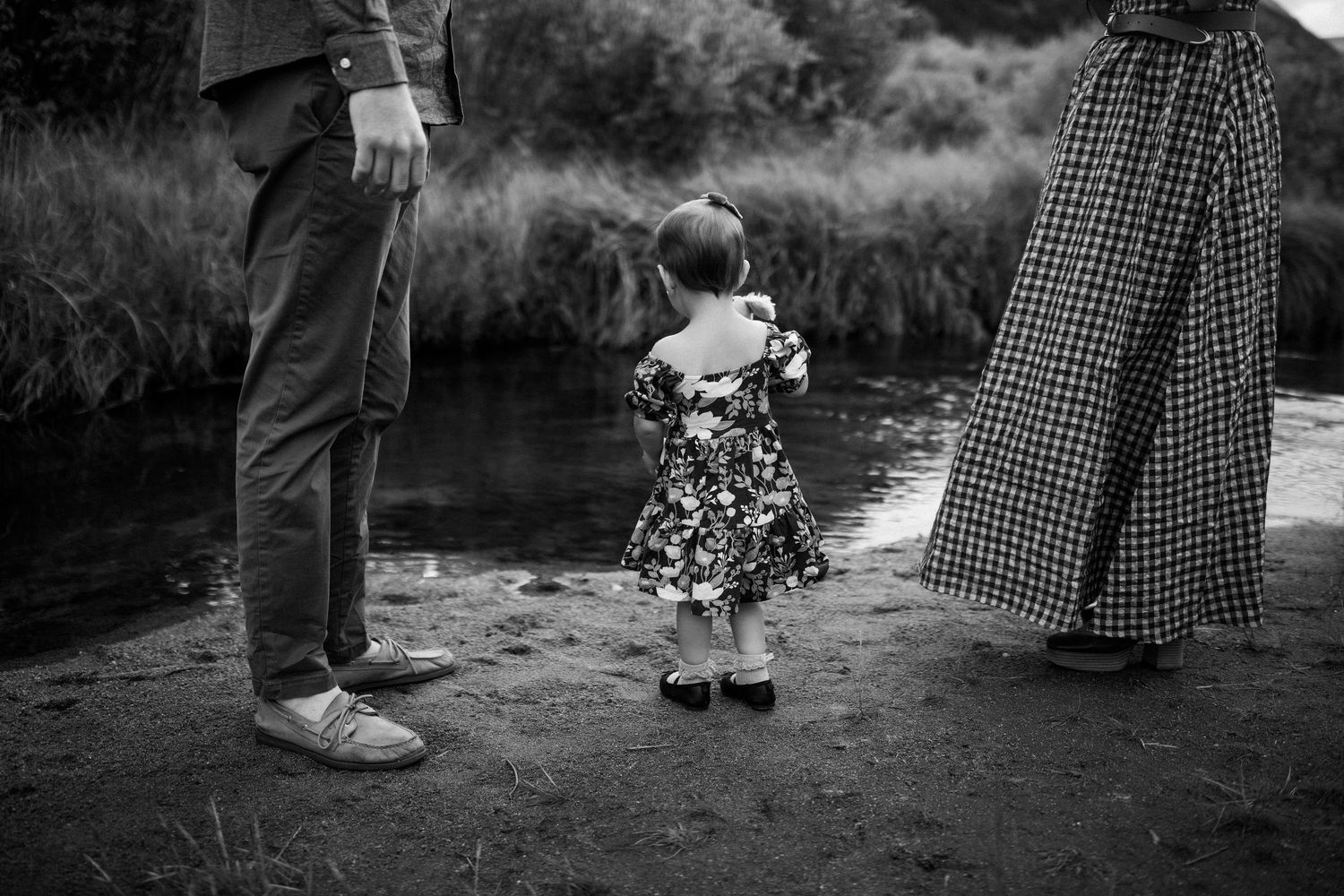 Black and white photo of a small child in a floral dress standing between two adults near a river bank.