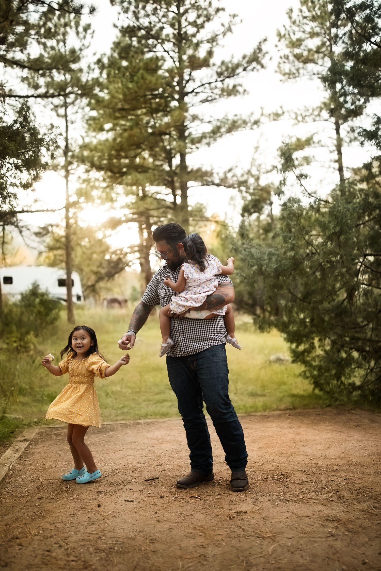 Family playing and dancing together on dirt path surrounded by pine trees at sunset.