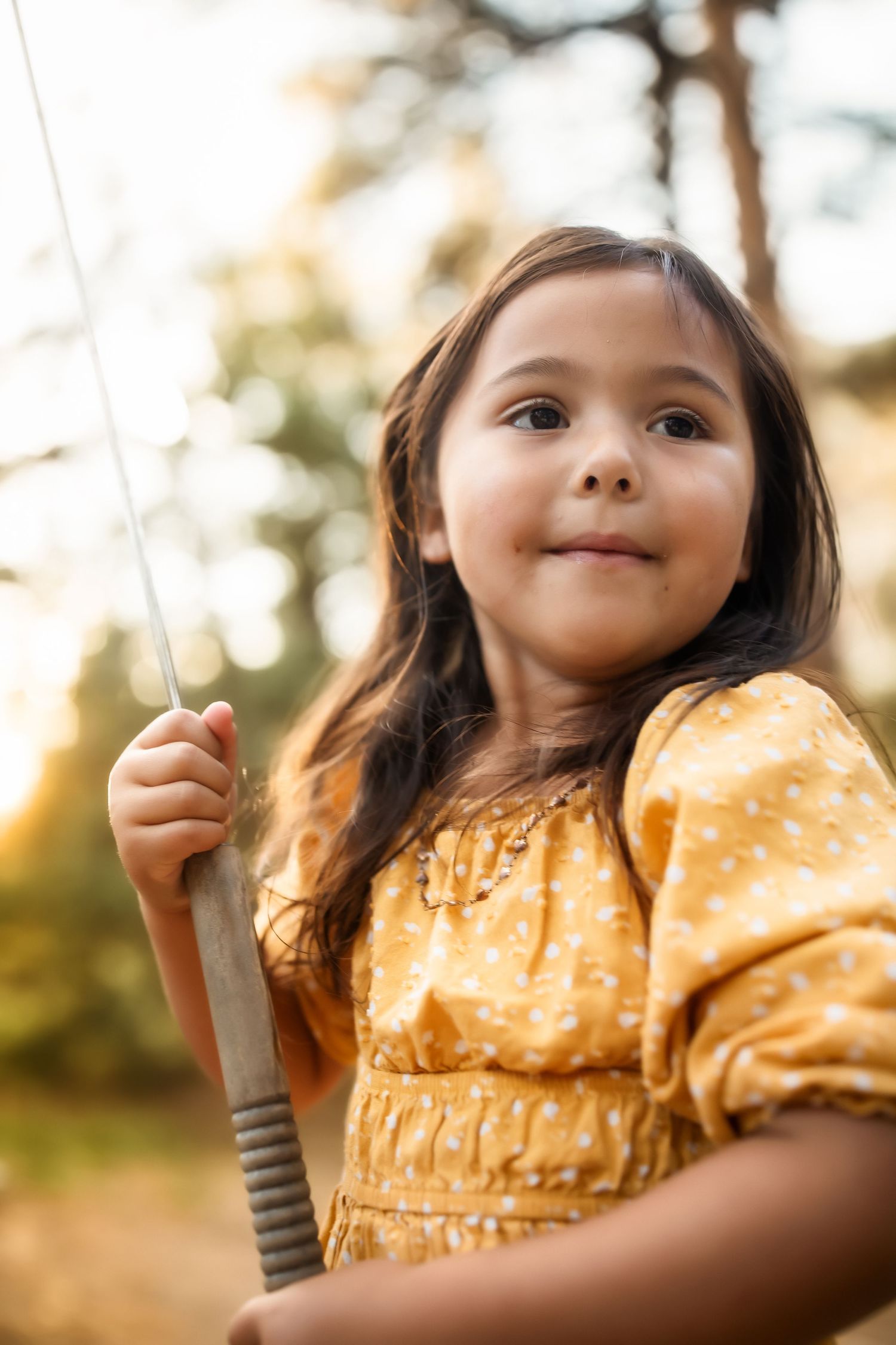 Child on swing wearing yellow dress smiling during golden hour photography session.
