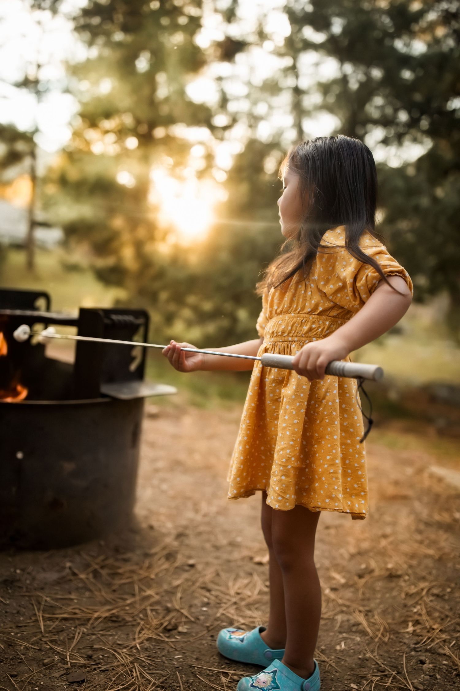 Child in yellow dress holding marshmallow roasting stick near campfire at sunset.