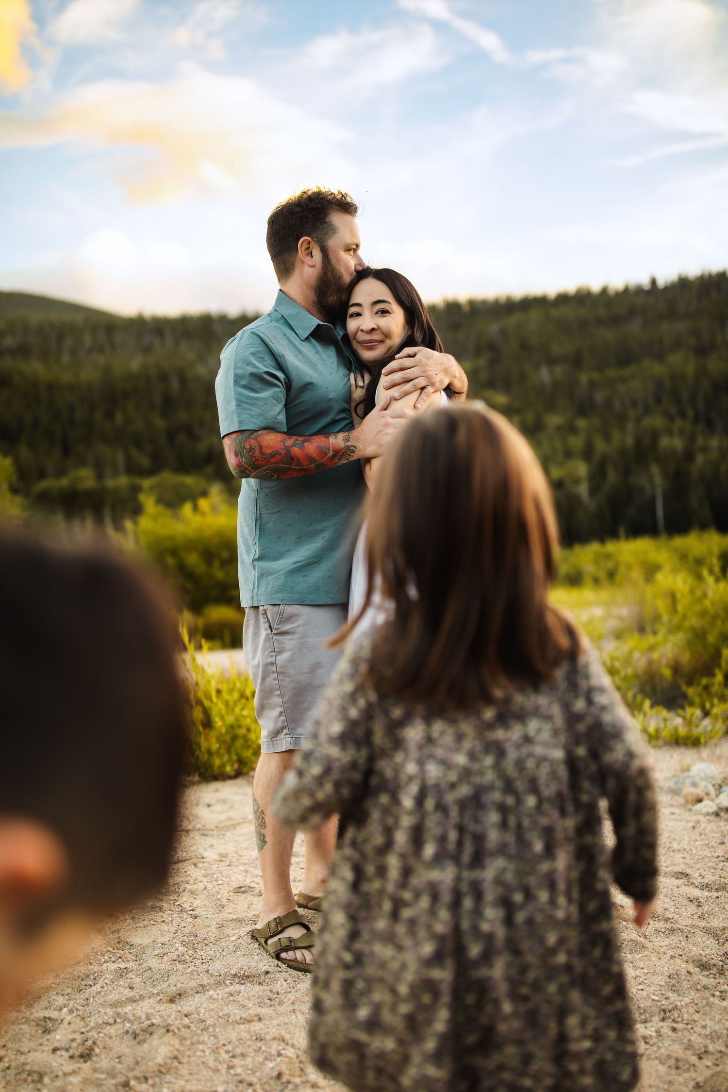 A couple embraces on a scenic mountain trail during golden hour with lush forest and hills in the background.
