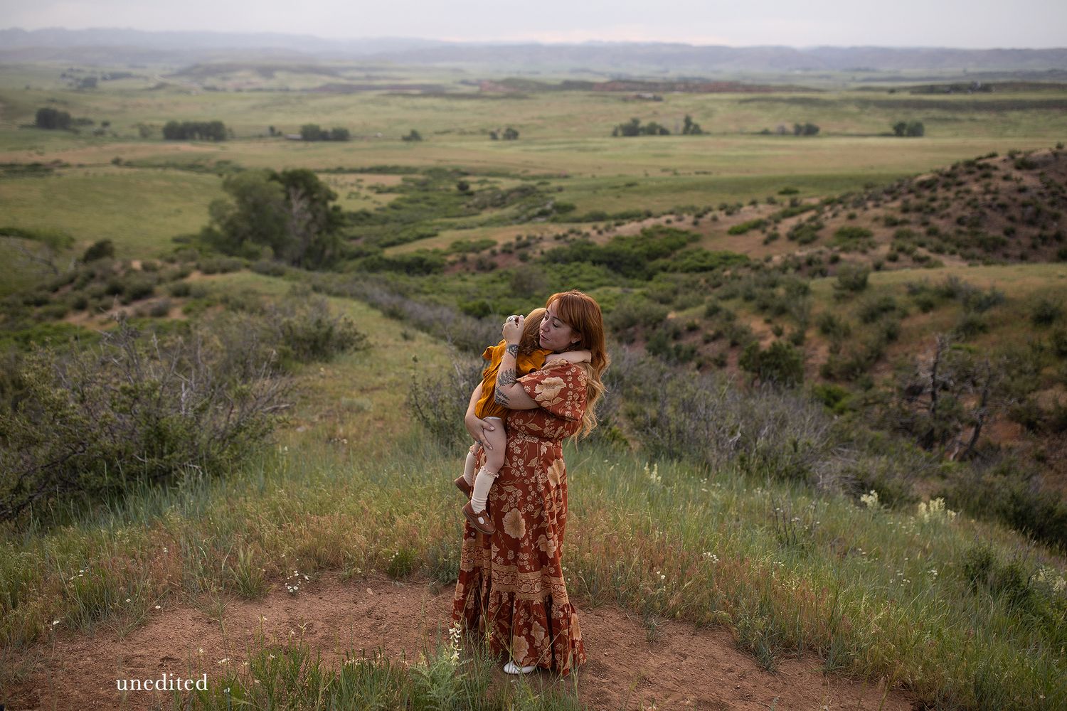 A couple embraces on a hilltop overlooking vast green plains and rolling hills during golden hour.