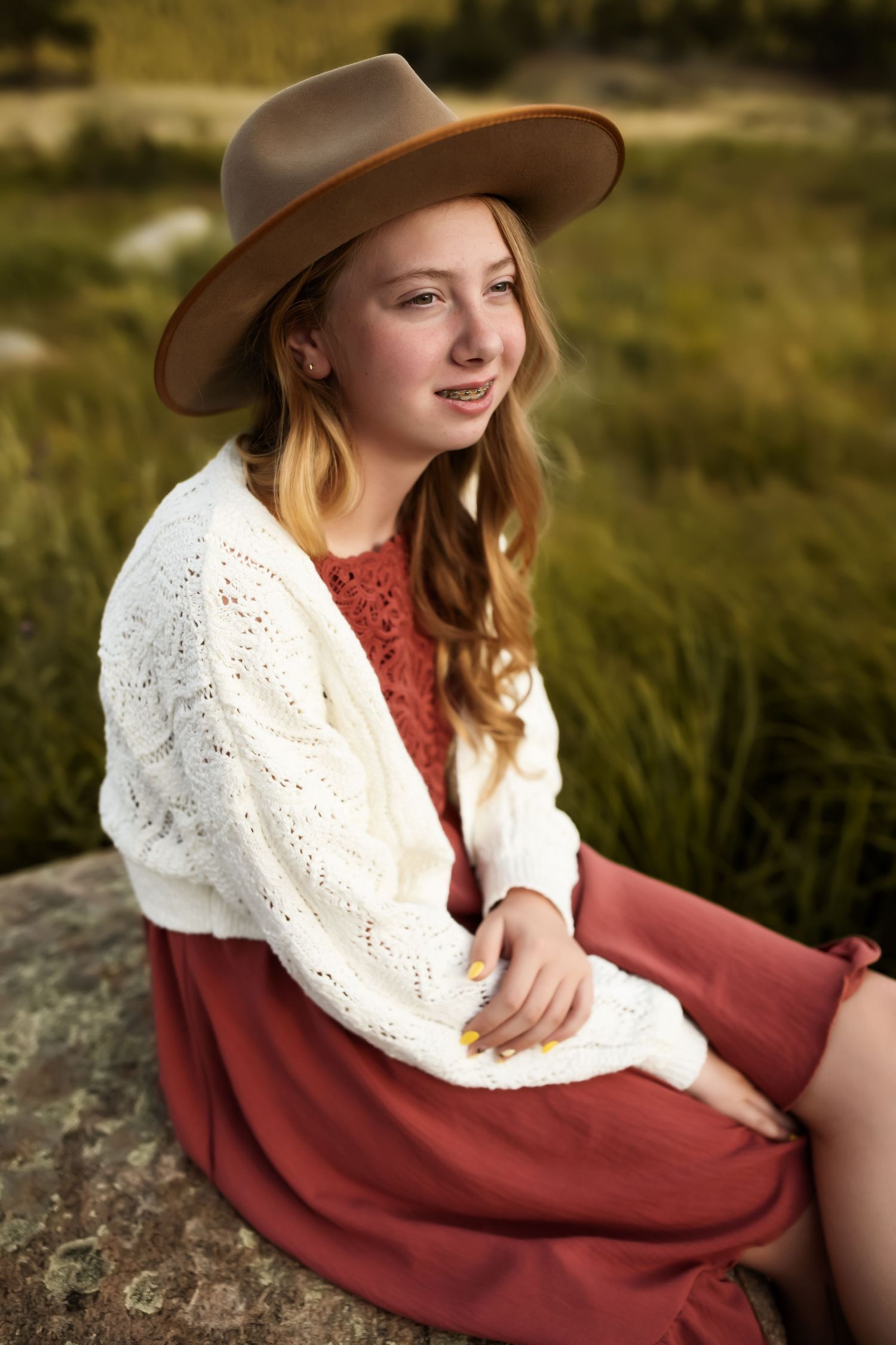 Person in a brown hat and burgundy dress sitting on rocks in a rustic outdoor setting.