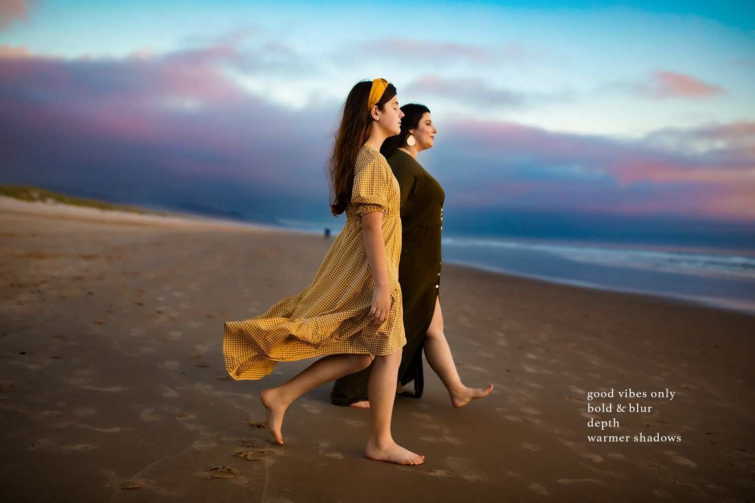 Couple walking barefoot along beach at sunset with flowing dresses and dramatic sky.
