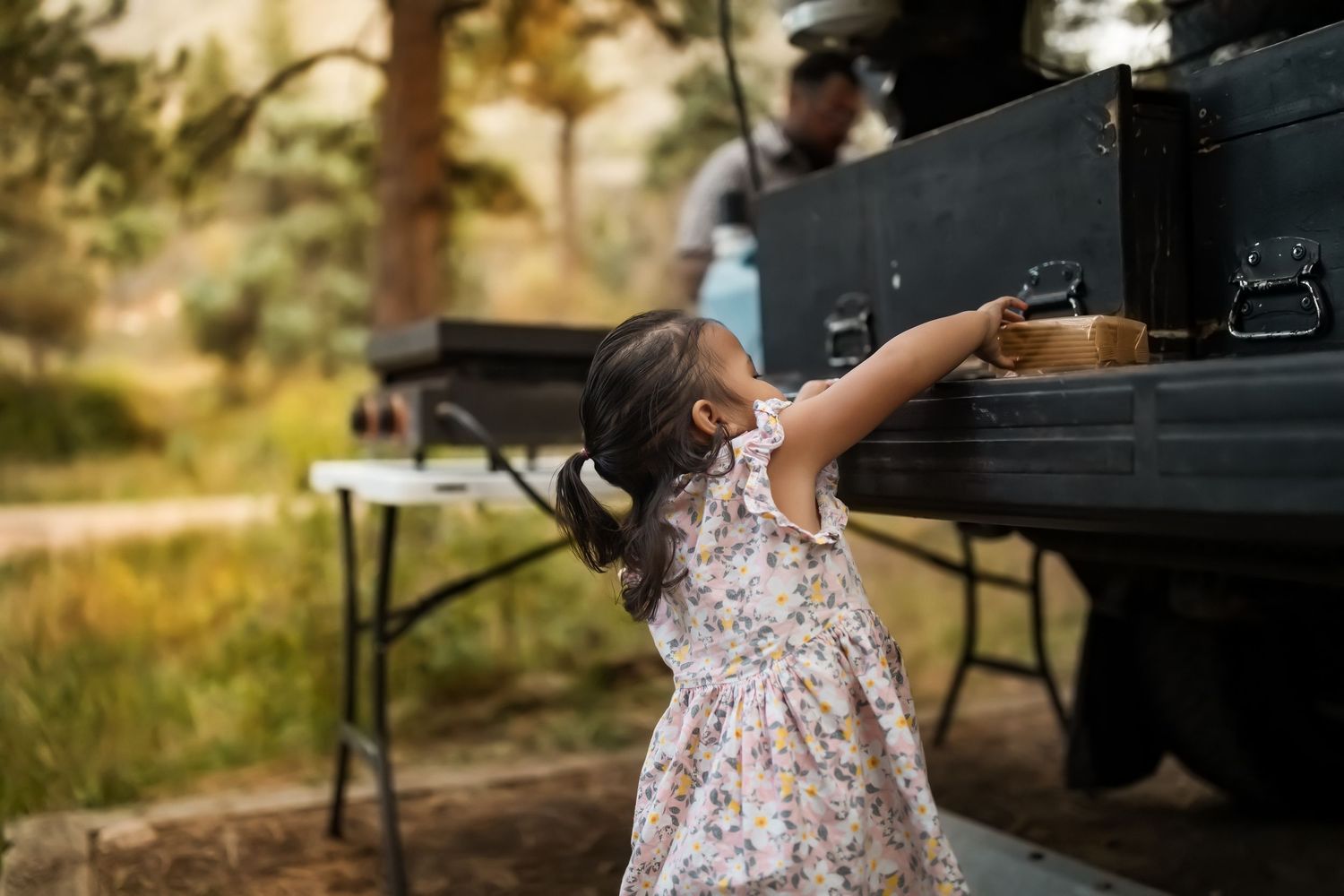 A child in a floral dress explores outdoors while adults sit at picnic tables in a sunny park setting.