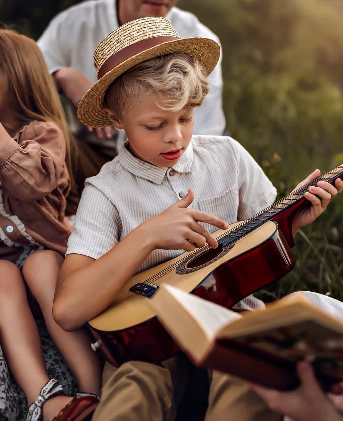 Young boy in straw hat playing acoustic guitar outdoors while family relaxes together in a sunny field.