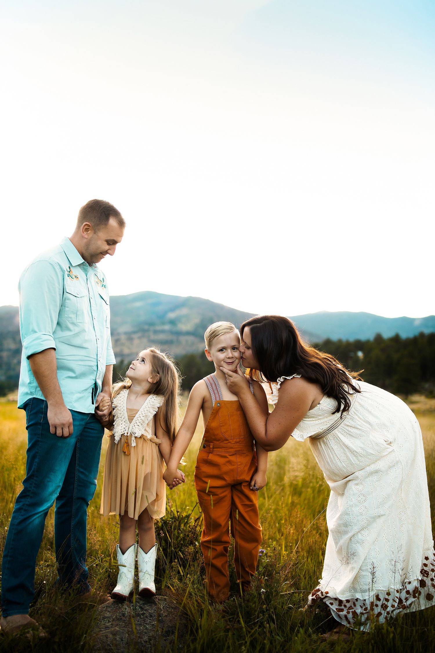Playful family moment captured in a sun-drenched field with mountains in the background.