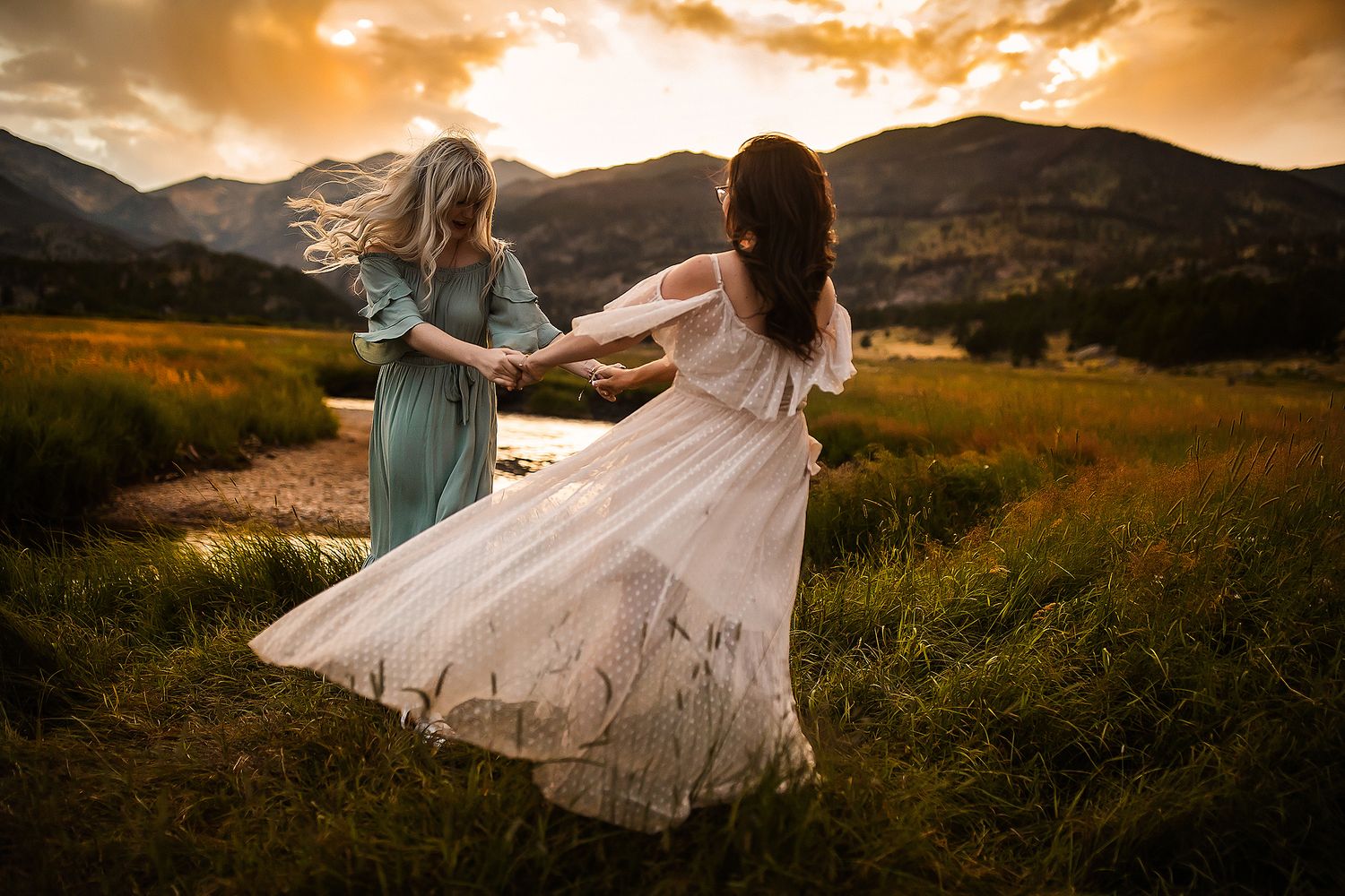 Two people in flowing dresses dancing in a mountain meadow at sunset.