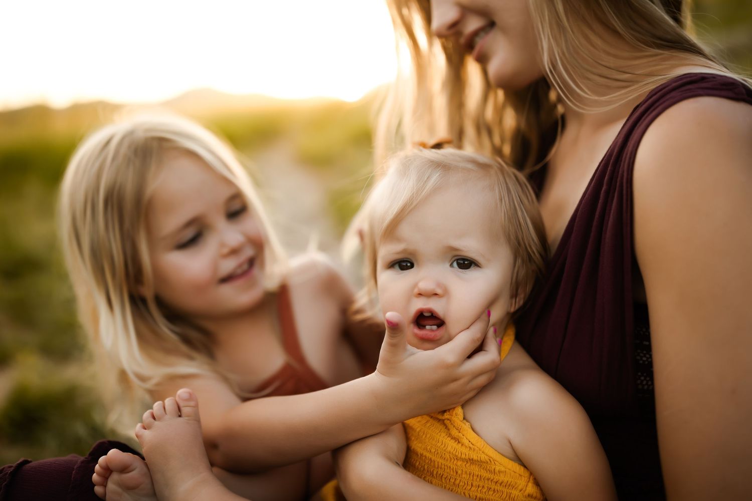 A mother holds two young blonde toddler girls outdoors in golden sunset light.