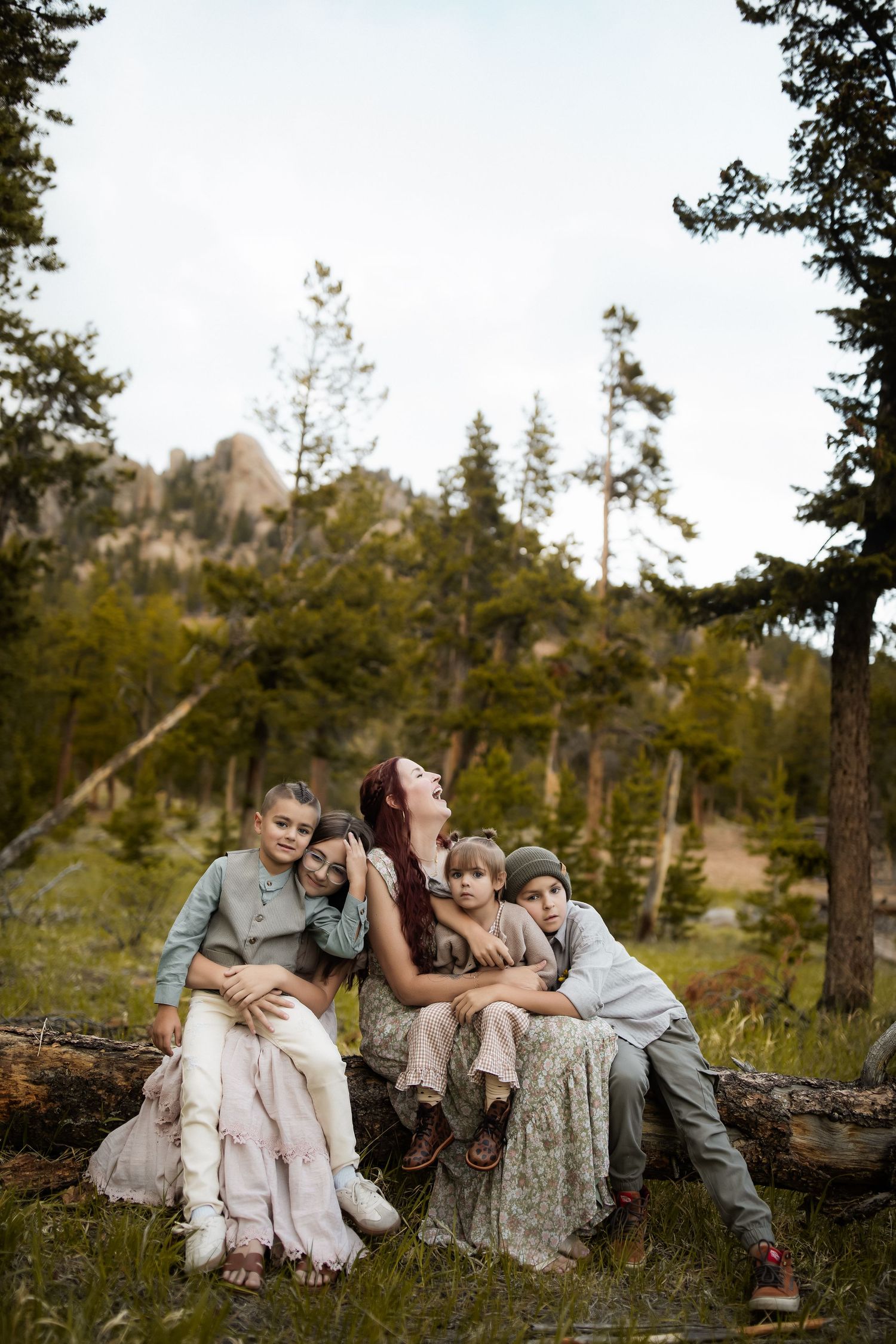 A mother sits on a log with three children in a forest meadow, surrounded by pine trees and rocky mountains.
