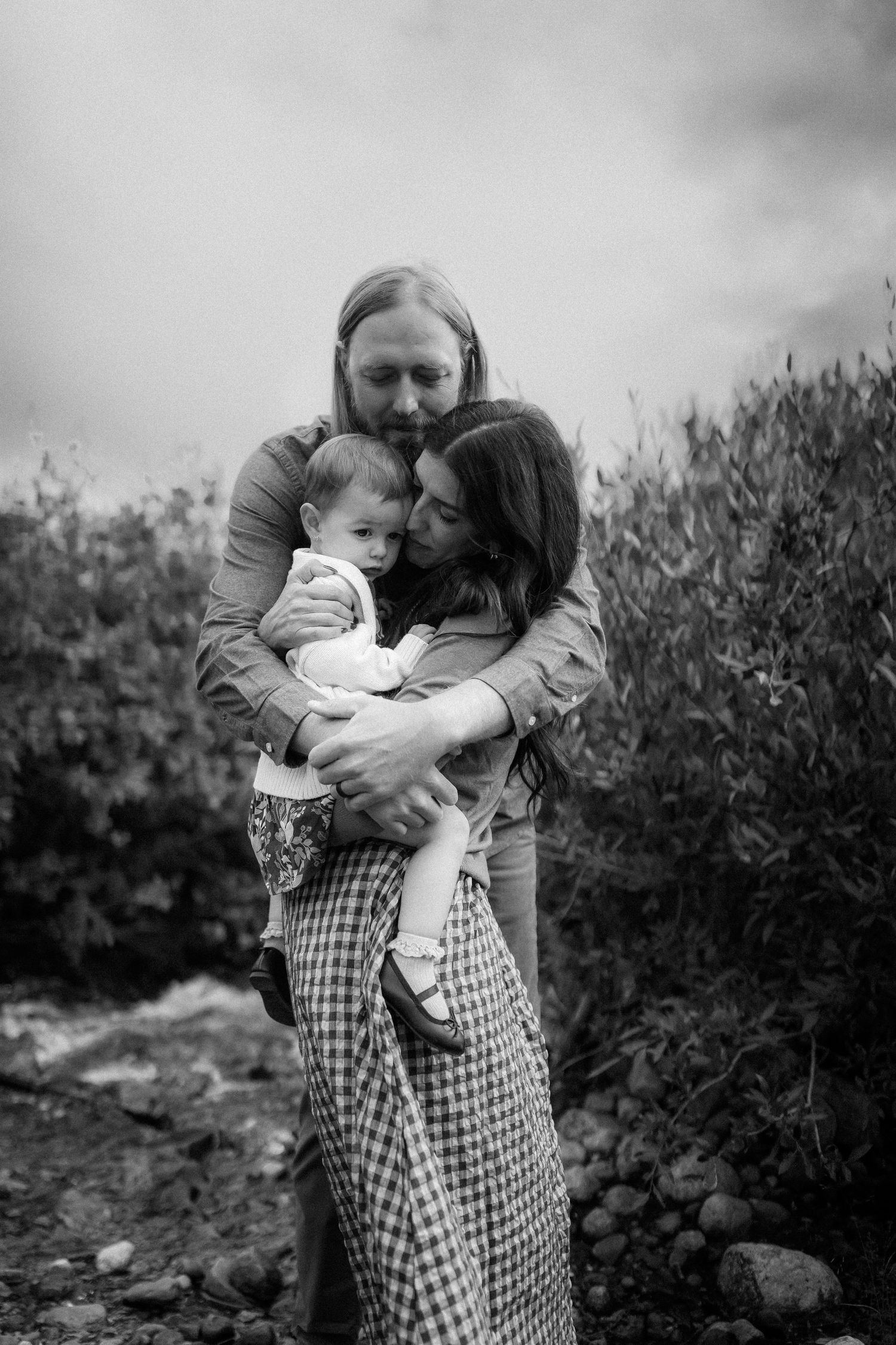 A black and white portrait captures an intimate family embrace outdoors.