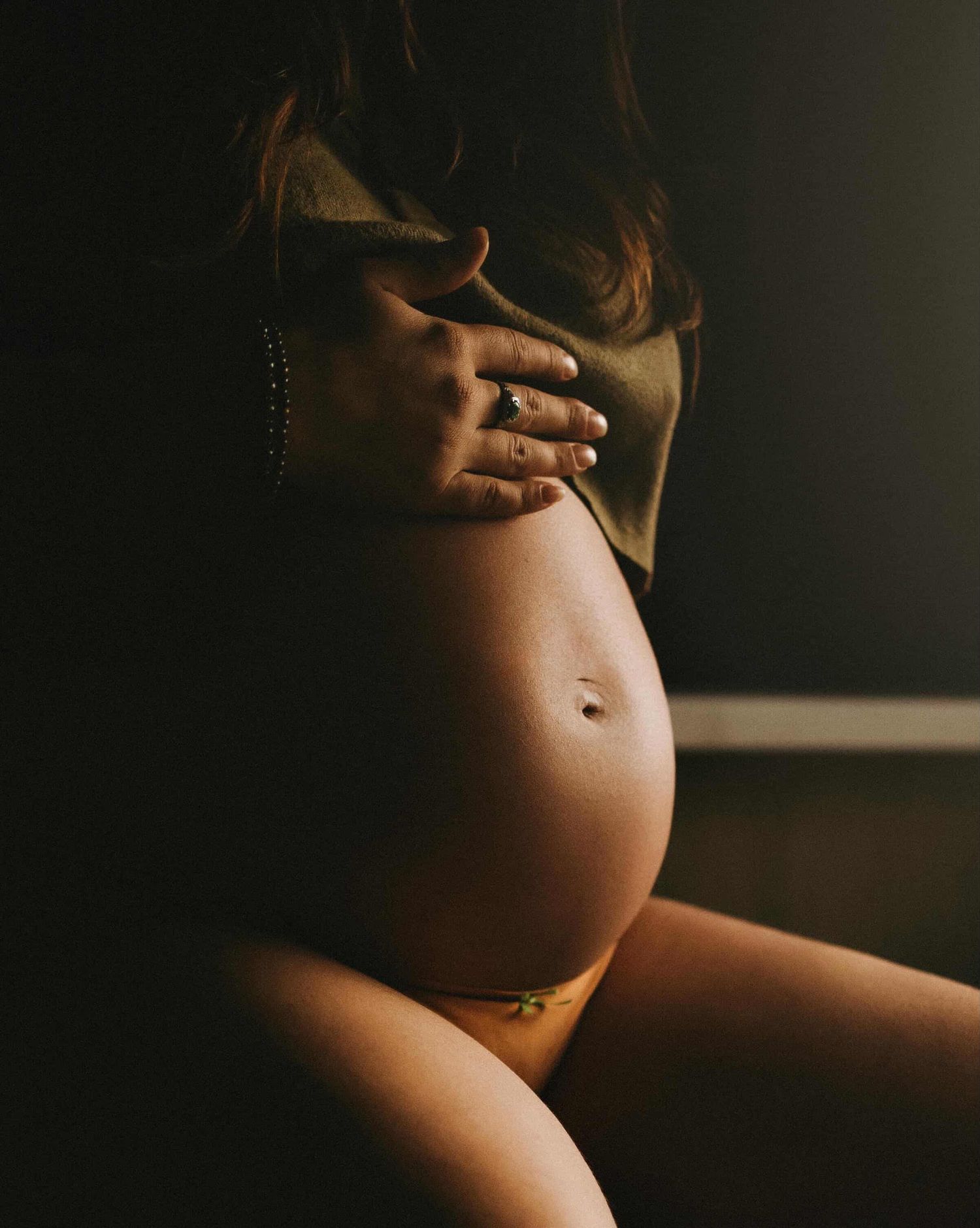 Close-up of a pregnant woman cradling her bare baby bump, wearing a green top, captured in moody, dramatic lighting.