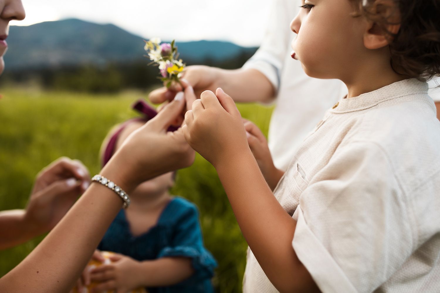 A person holds and blows on a delicate white dandelion flower in a sunlit outdoor setting.