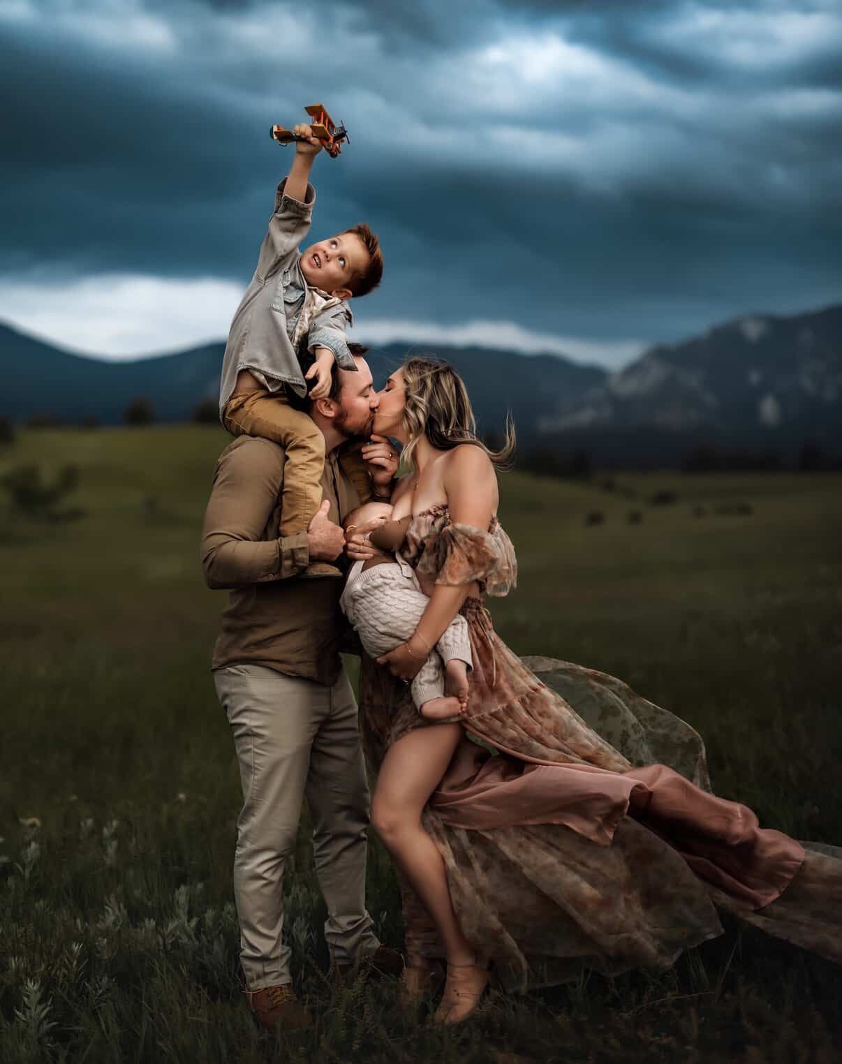 Family portrait in a mountain meadow under dramatic stormy skies, parents kissing while children play with a toy airplane.