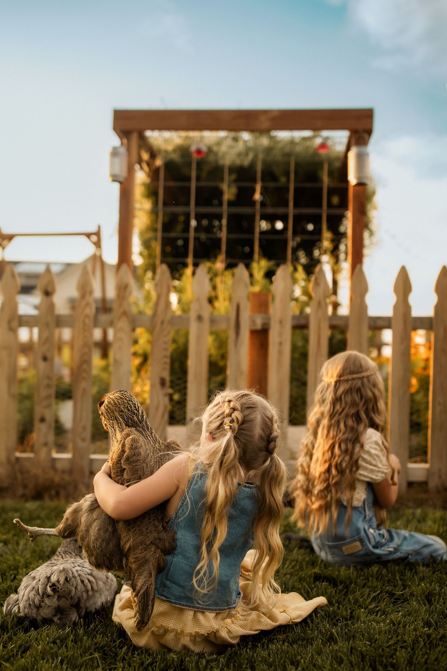 Two girls with long hair sit on grass facing a wooden fence and floral arch, one holding a chicken.
