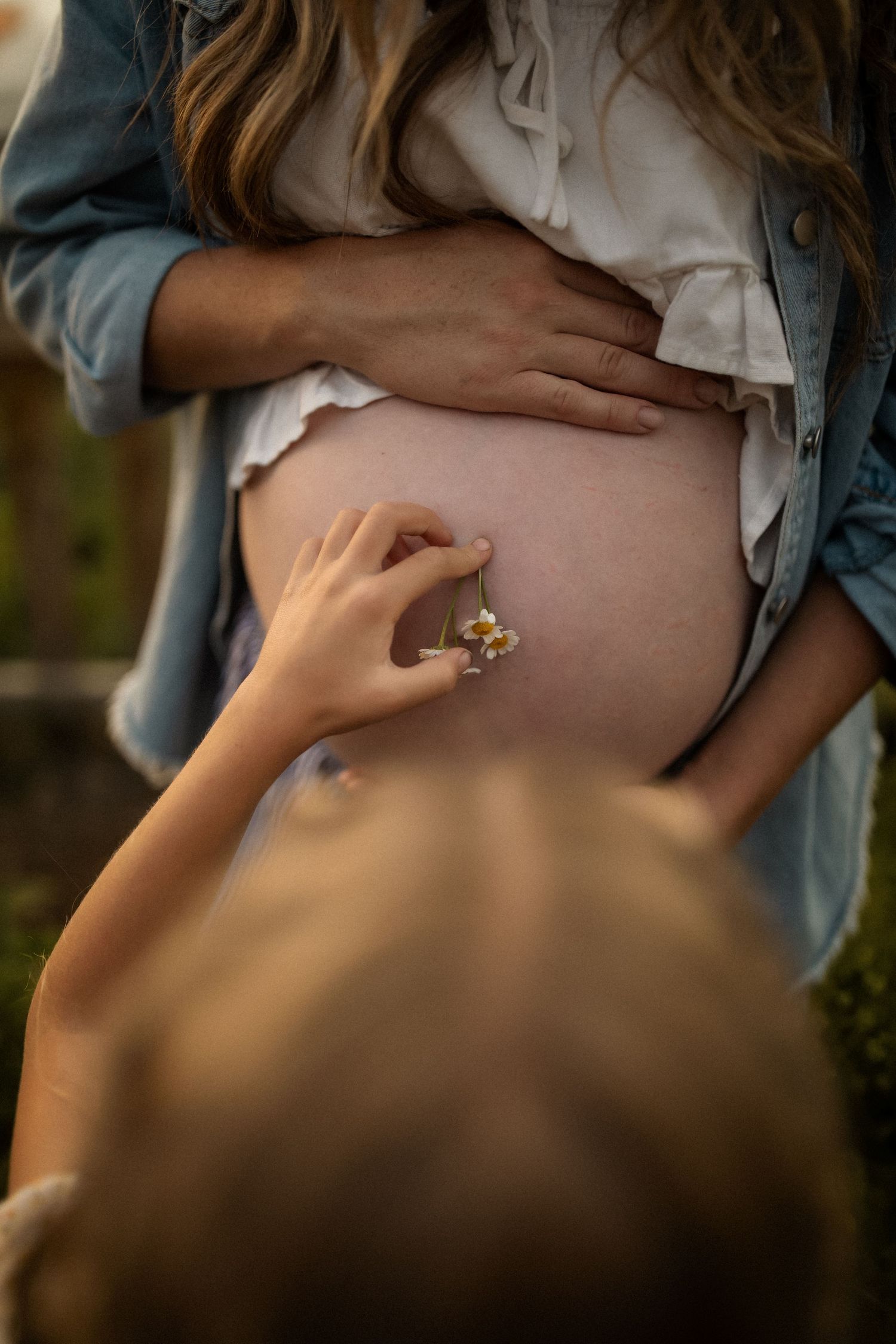 A small hand reaches out to touch a pregnant belly in a tender moment captured in soft lighting.