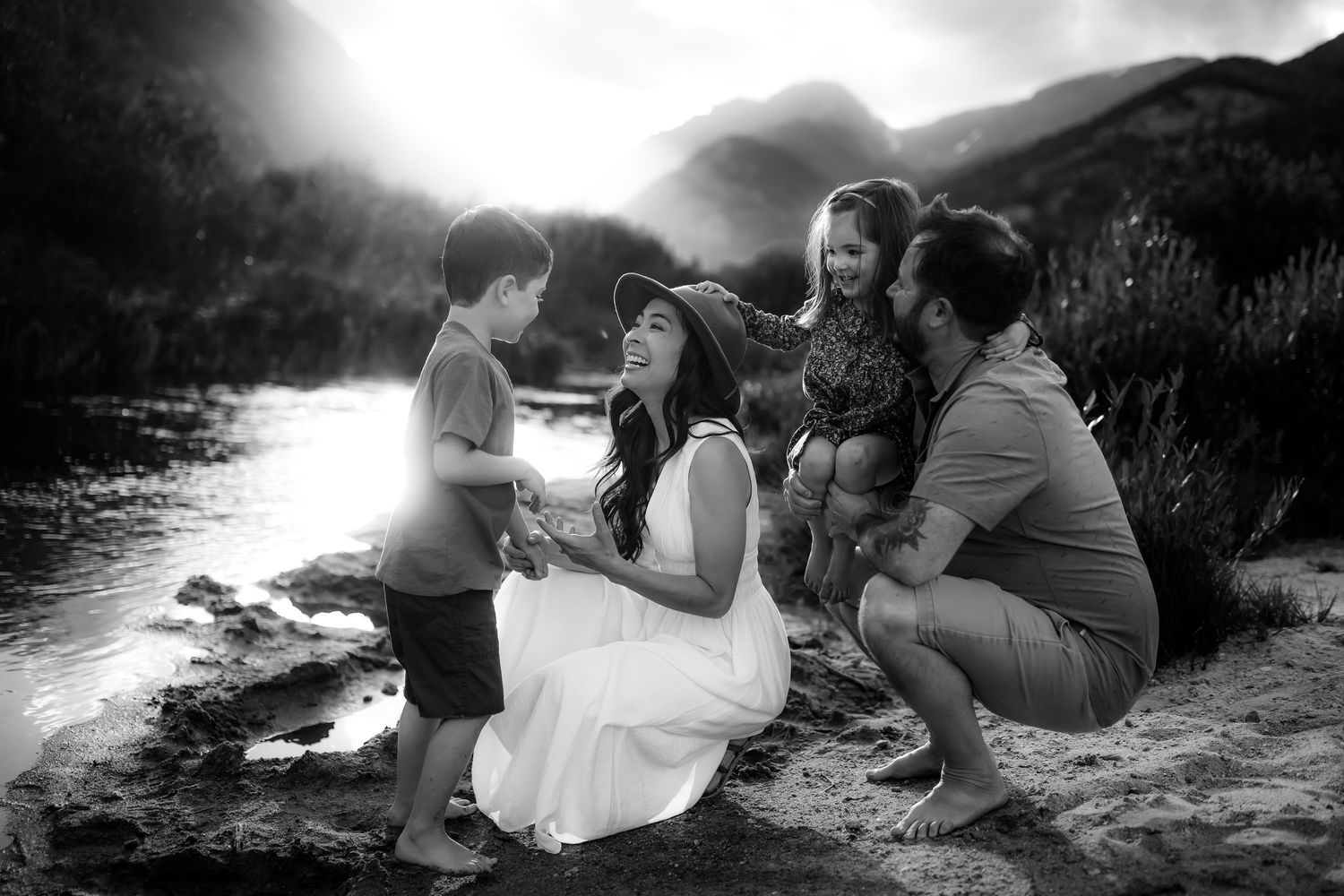 Black and white family photo of parents and two children sharing a joyful moment by a sunlit river.