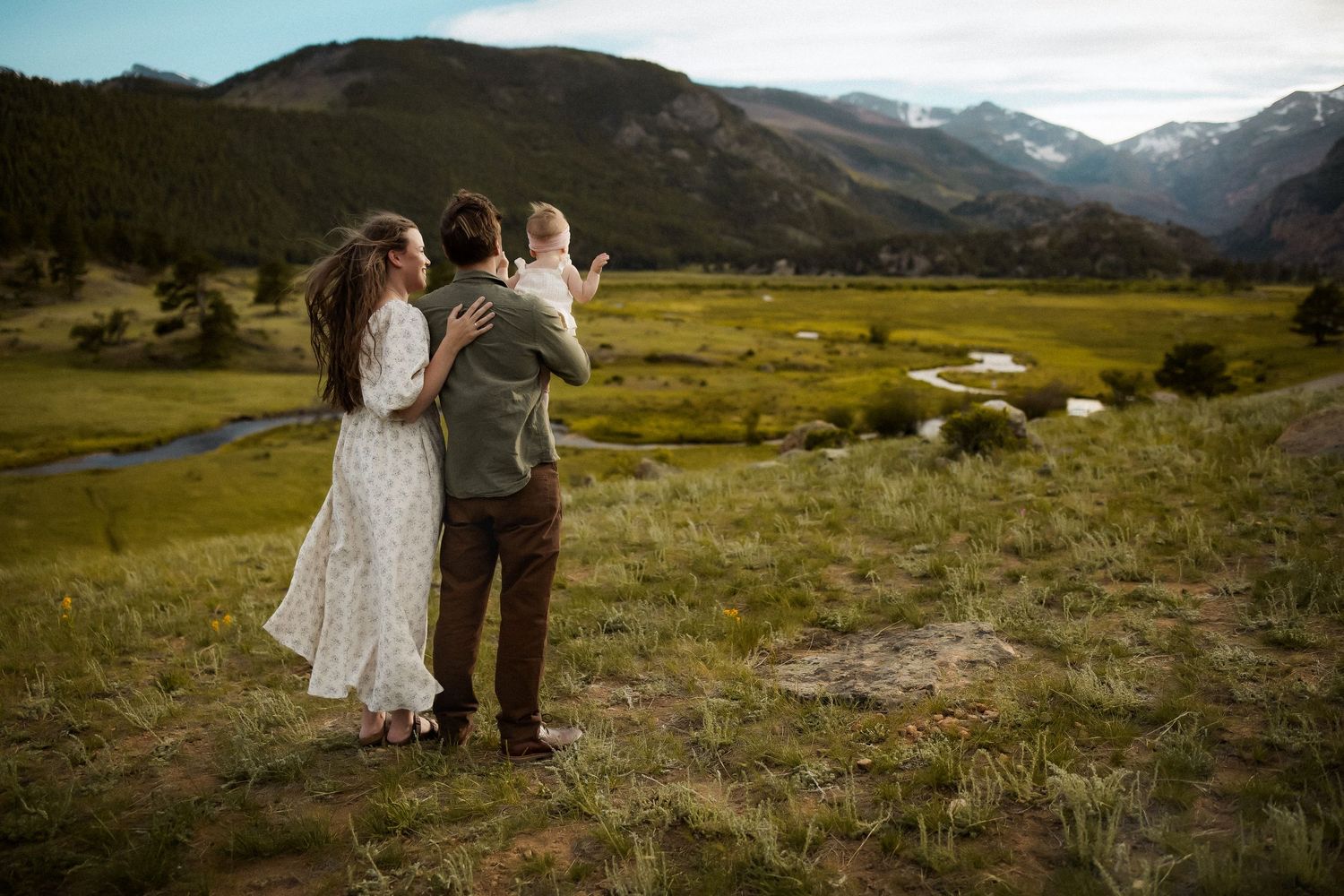 Family of three standing together in a lush green mountain valley landscape with scenic peaks in the background.