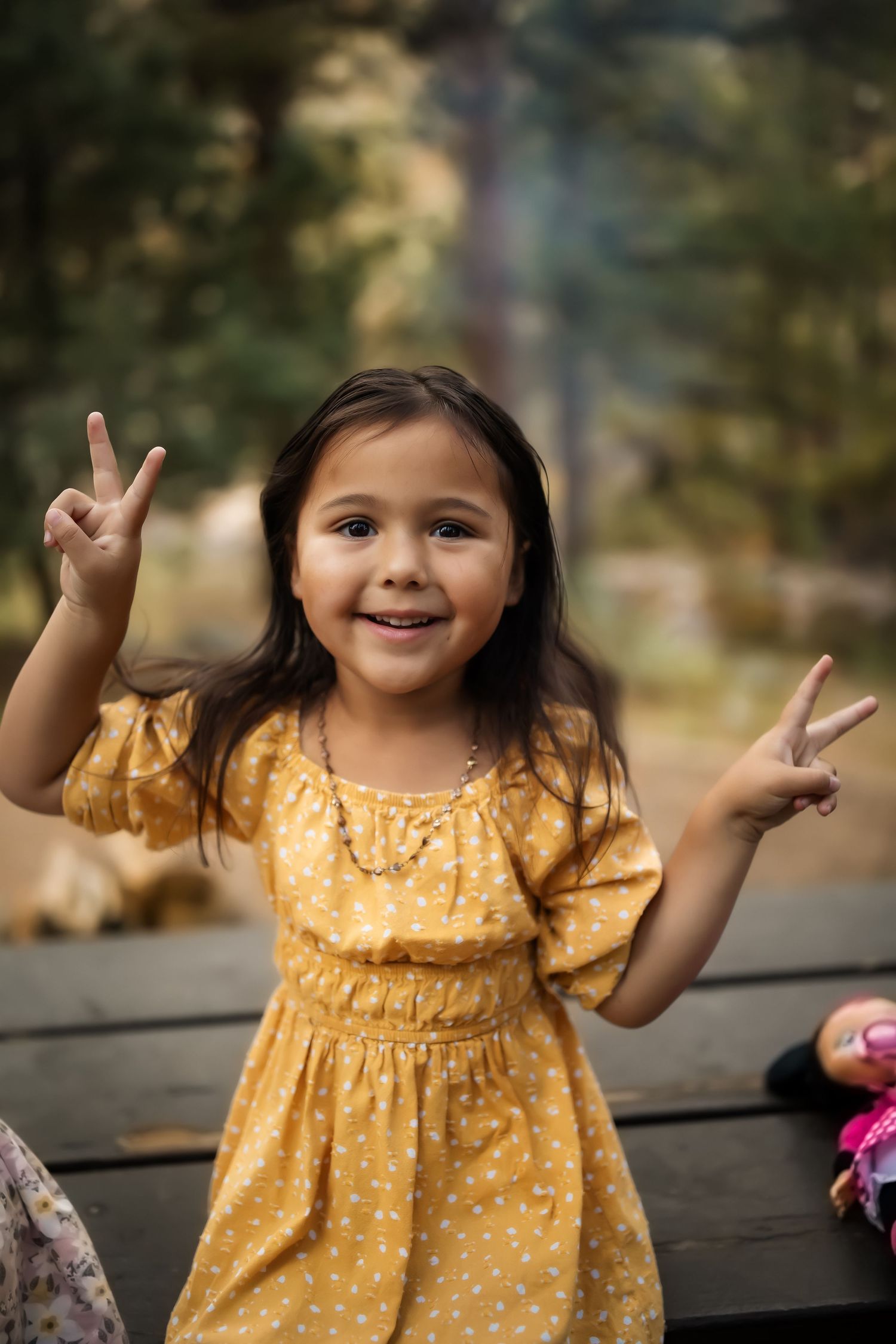 Young child in yellow polka dot dress smiling and making peace signs while standing outdoors.