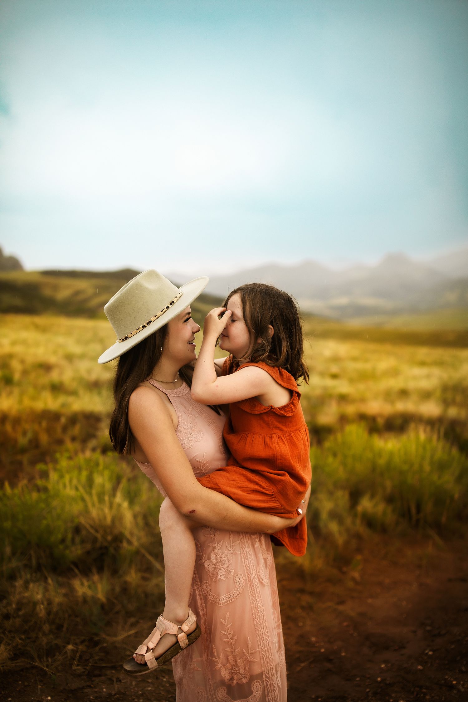 A mother in a pink dress and white hat holds her young daughter in an orange dress, sharing a tender kiss in a green meadow.