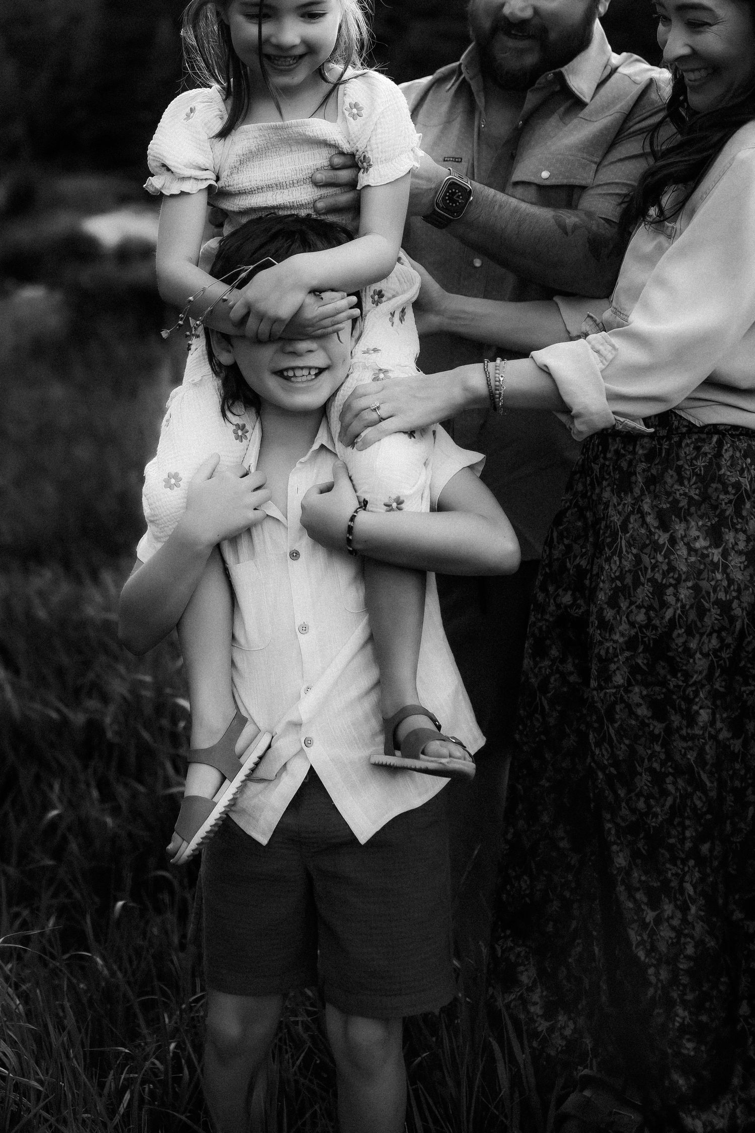 Black and white family photo of boy with girl on shoulders, parents laughing outdoors in grassy field.
