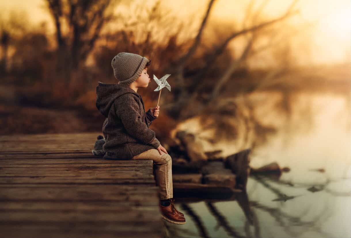 A young child sits on a wooden dock at sunset, holding a pinwheel toy beside a calm autumn lake.