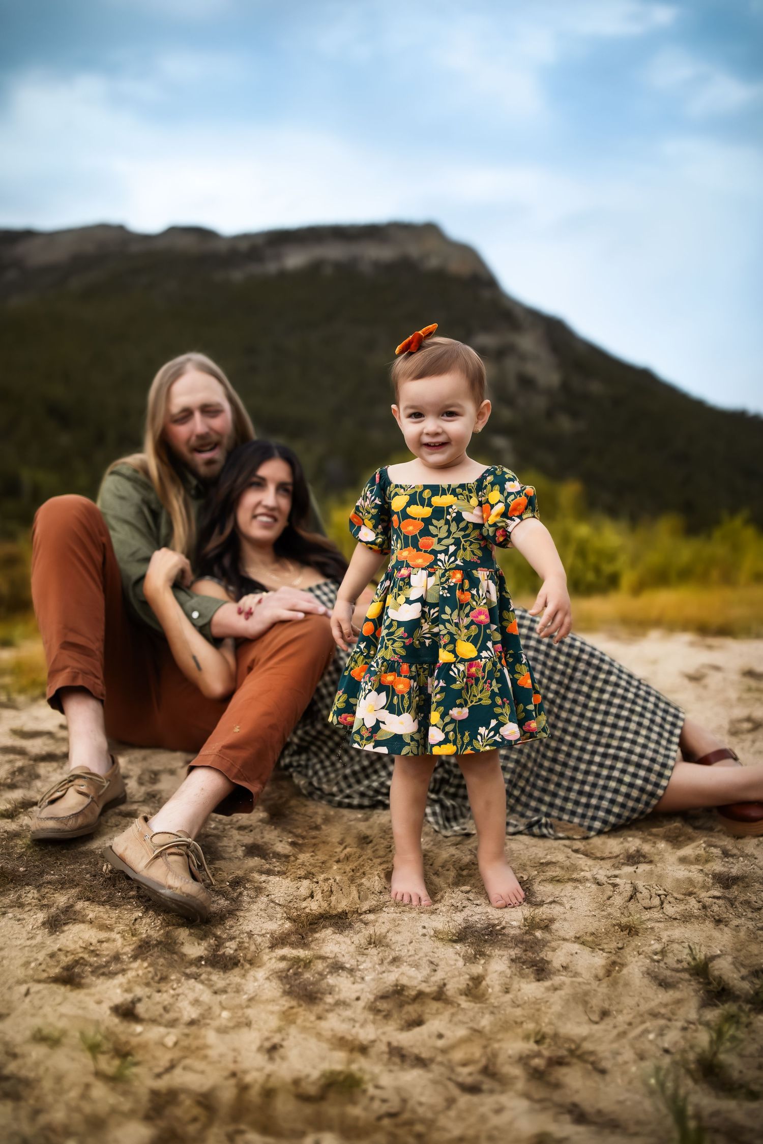 A family relaxes on a picnic blanket near mountains with their toddler in a floral dress standing in front.