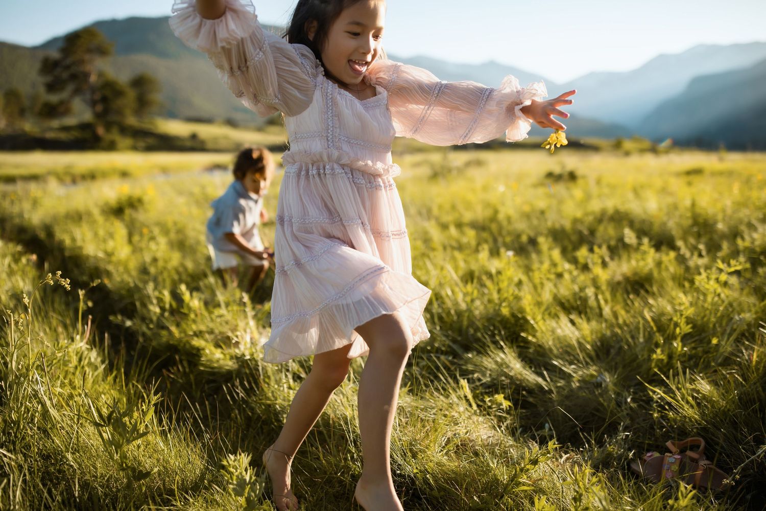 Child in white dress runs playfully through a sunlit meadow with mountains in the background.
