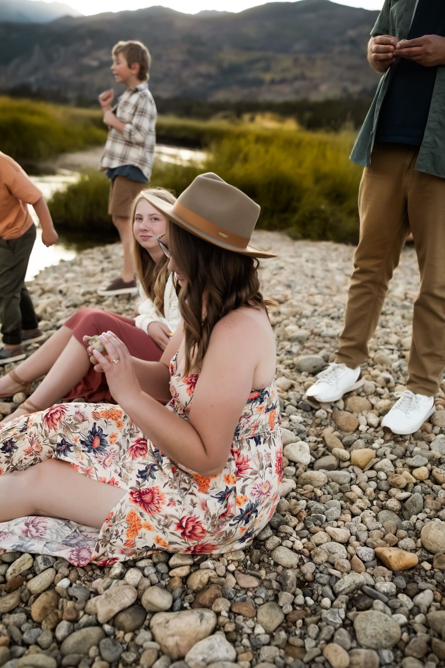 People relaxing on rocky riverbank with mountains in background during outdoor gathering.