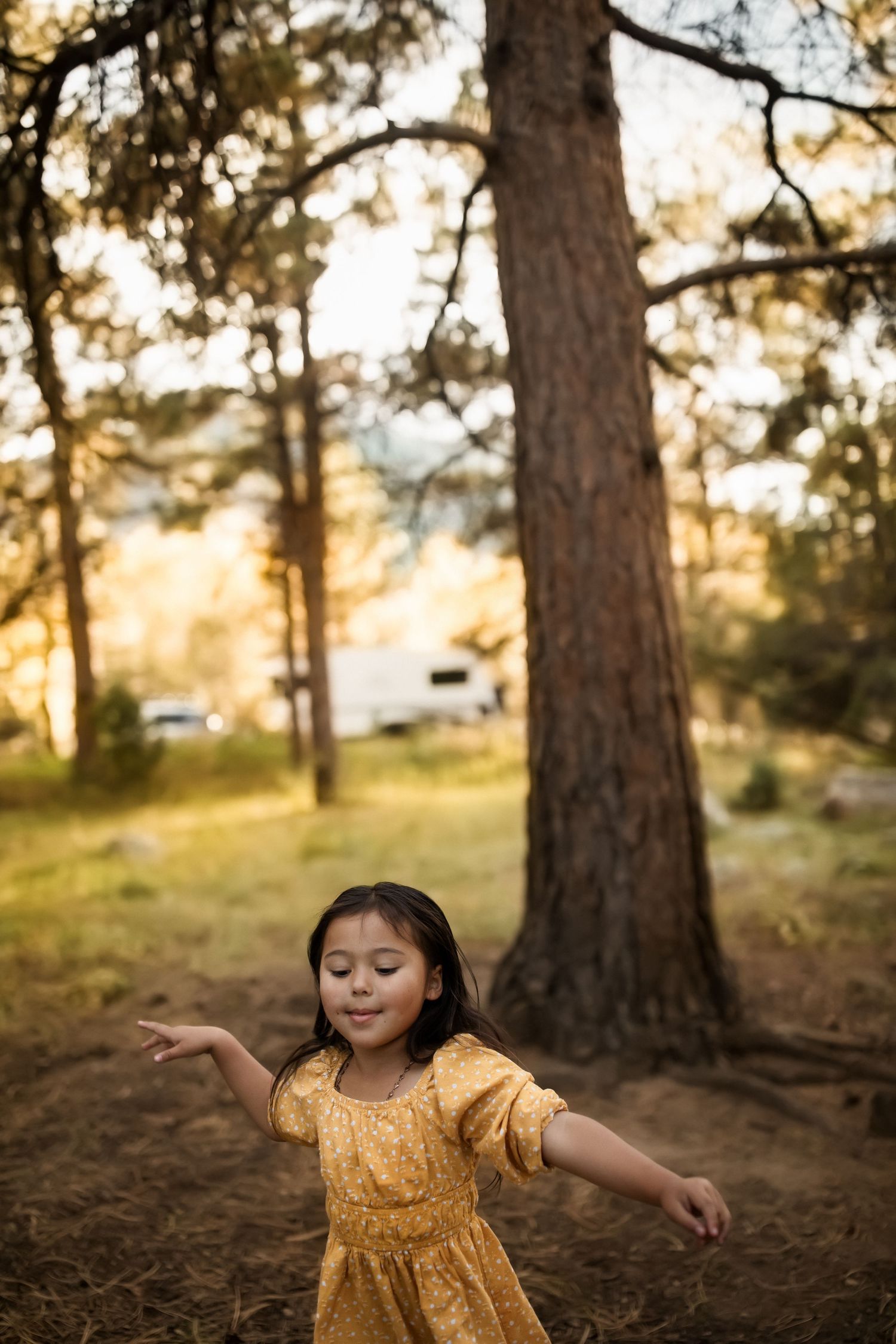 Child in yellow dress dancing and twirling among tall pine trees at sunset.