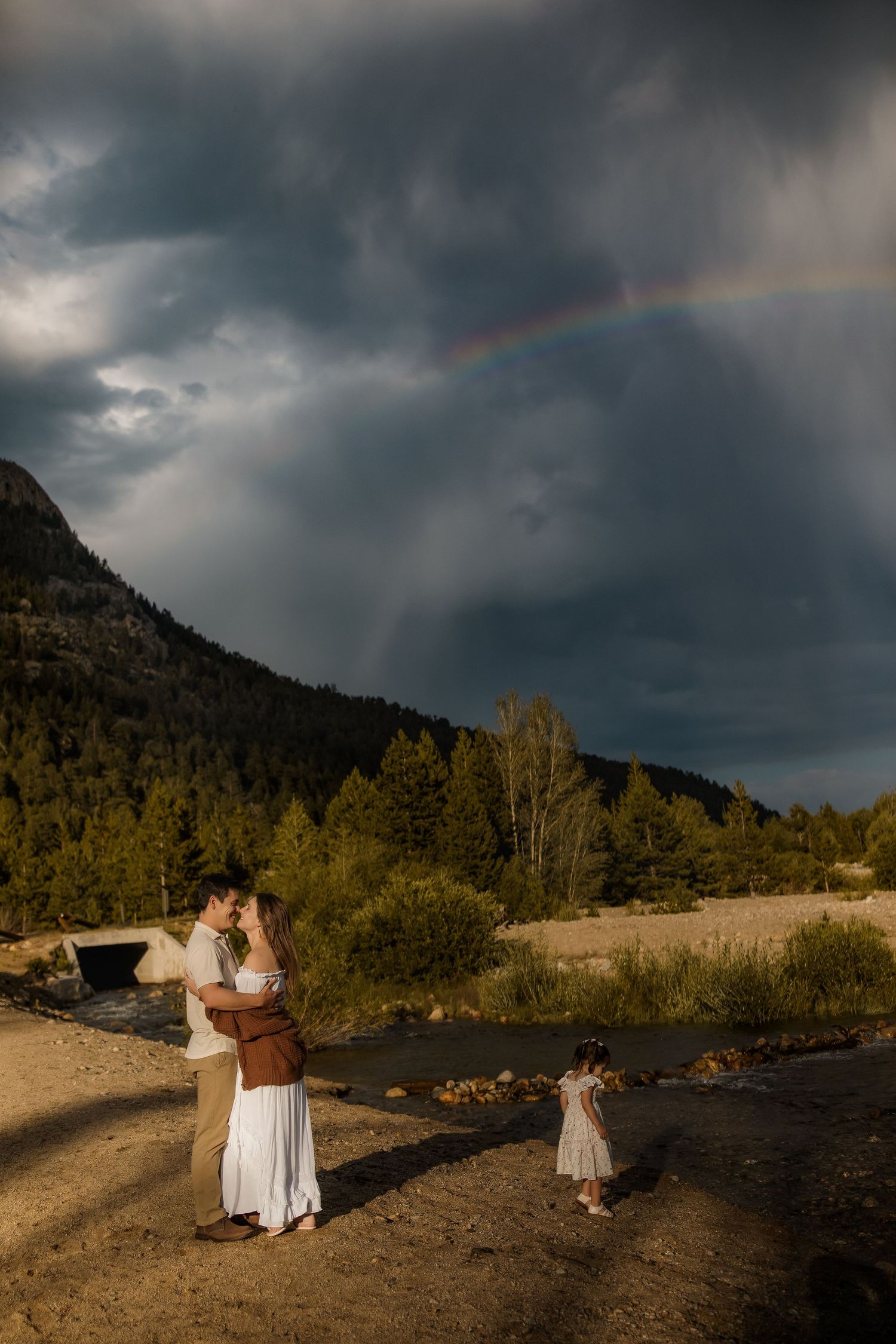 Couple shares a kiss near a mountain stream with a rainbow over stormy skies, as a child plays nearby.