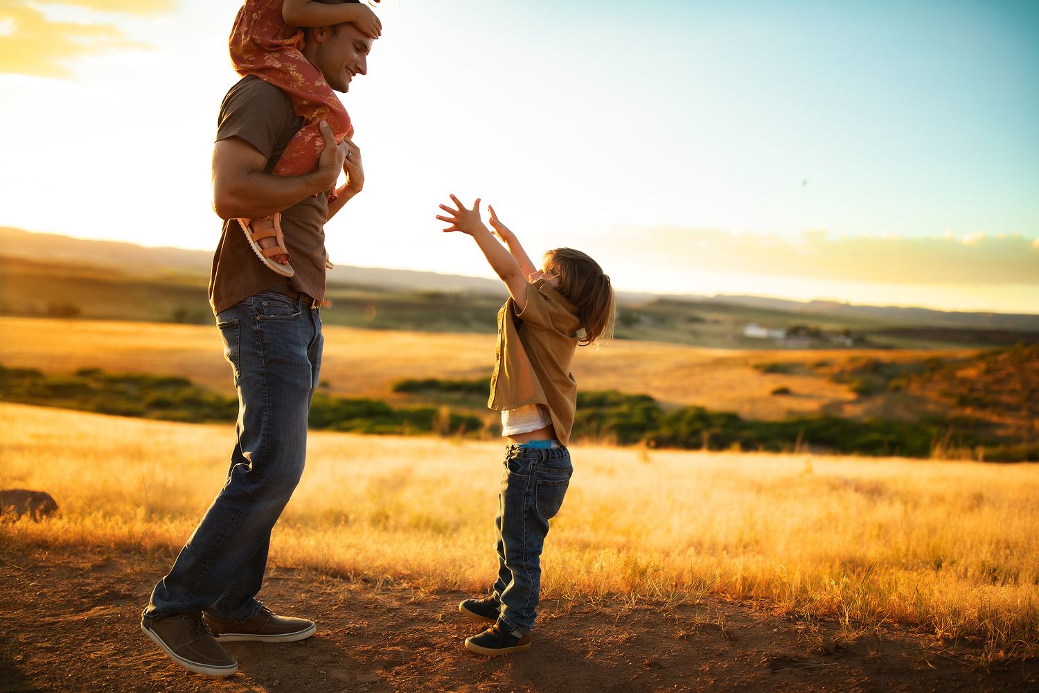 Two people stand close together in a golden wheat field at sunset with rolling hills in the background.