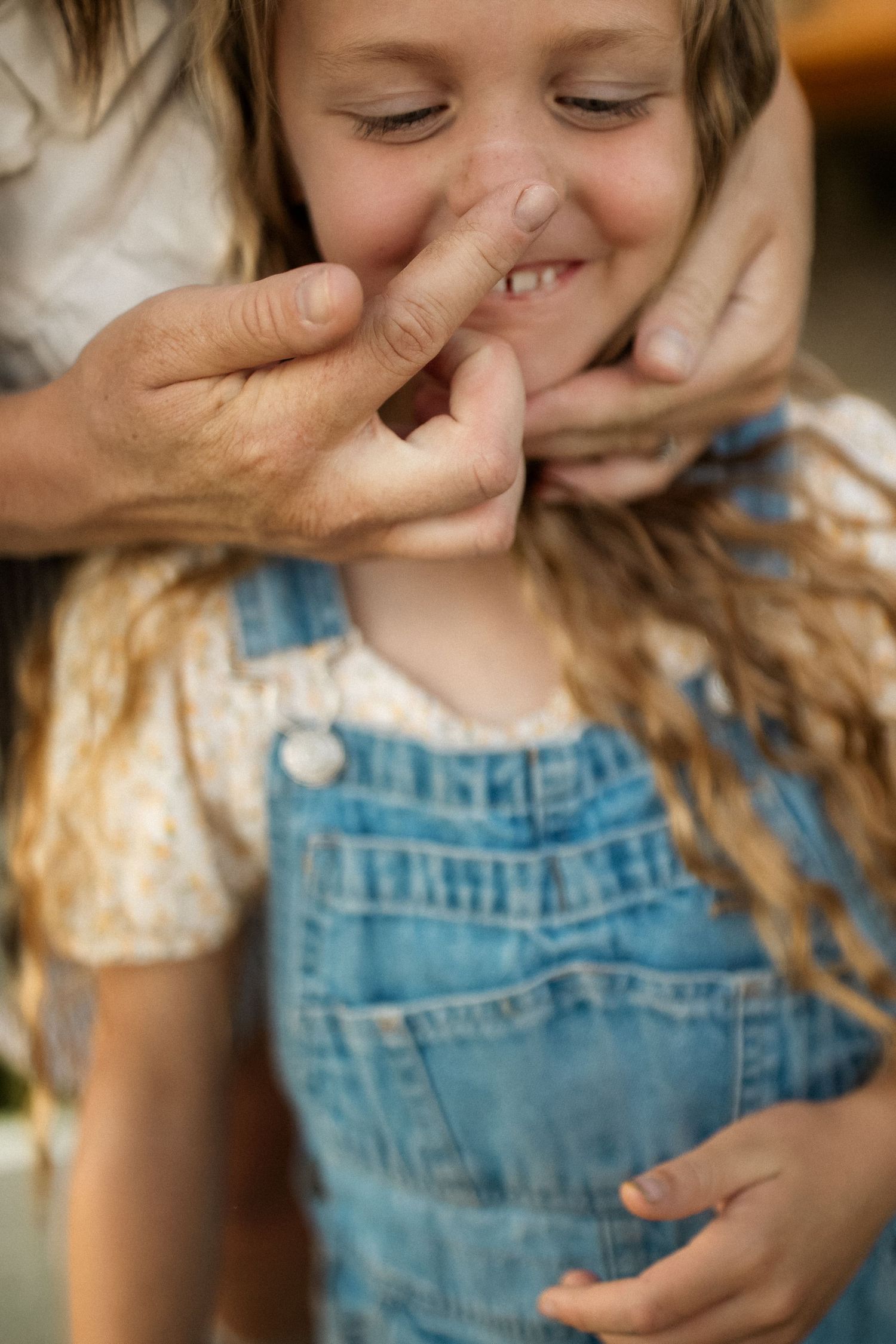 A playful moment captured as hands gently pinch a child's nose while wearing blue denim overalls.