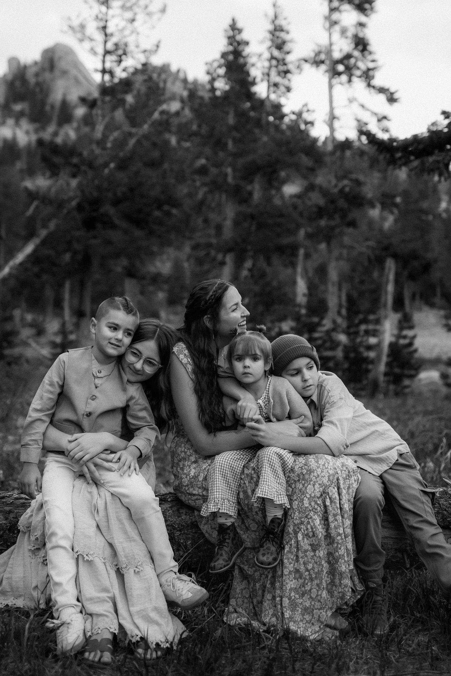 Black and white photo of a mother sitting outdoors with four children, surrounded by pine trees and rocky mountains.