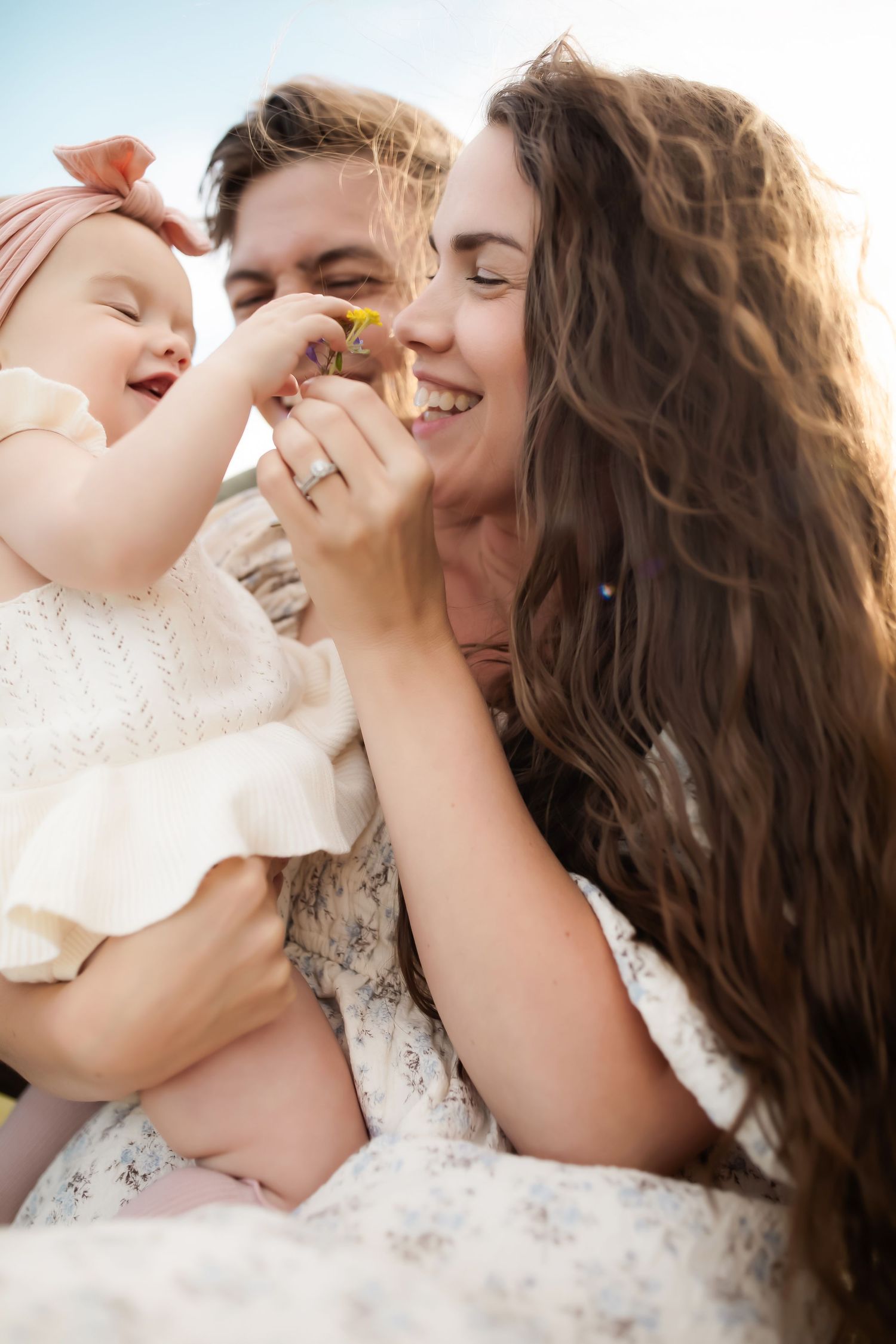 Happy young family with baby girl holding a flower outdoors in warm sunlight, sharing a joyful moment together.