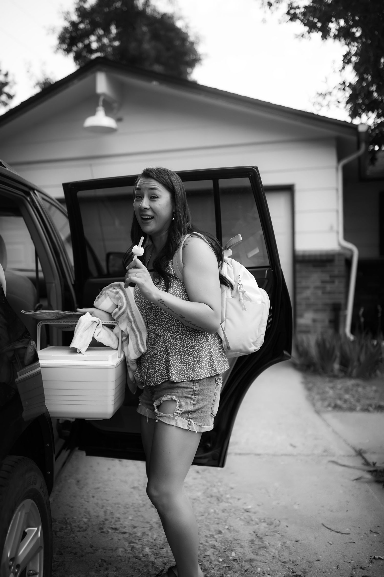 Black and white photo of someone leaning into a car parked outside a house.