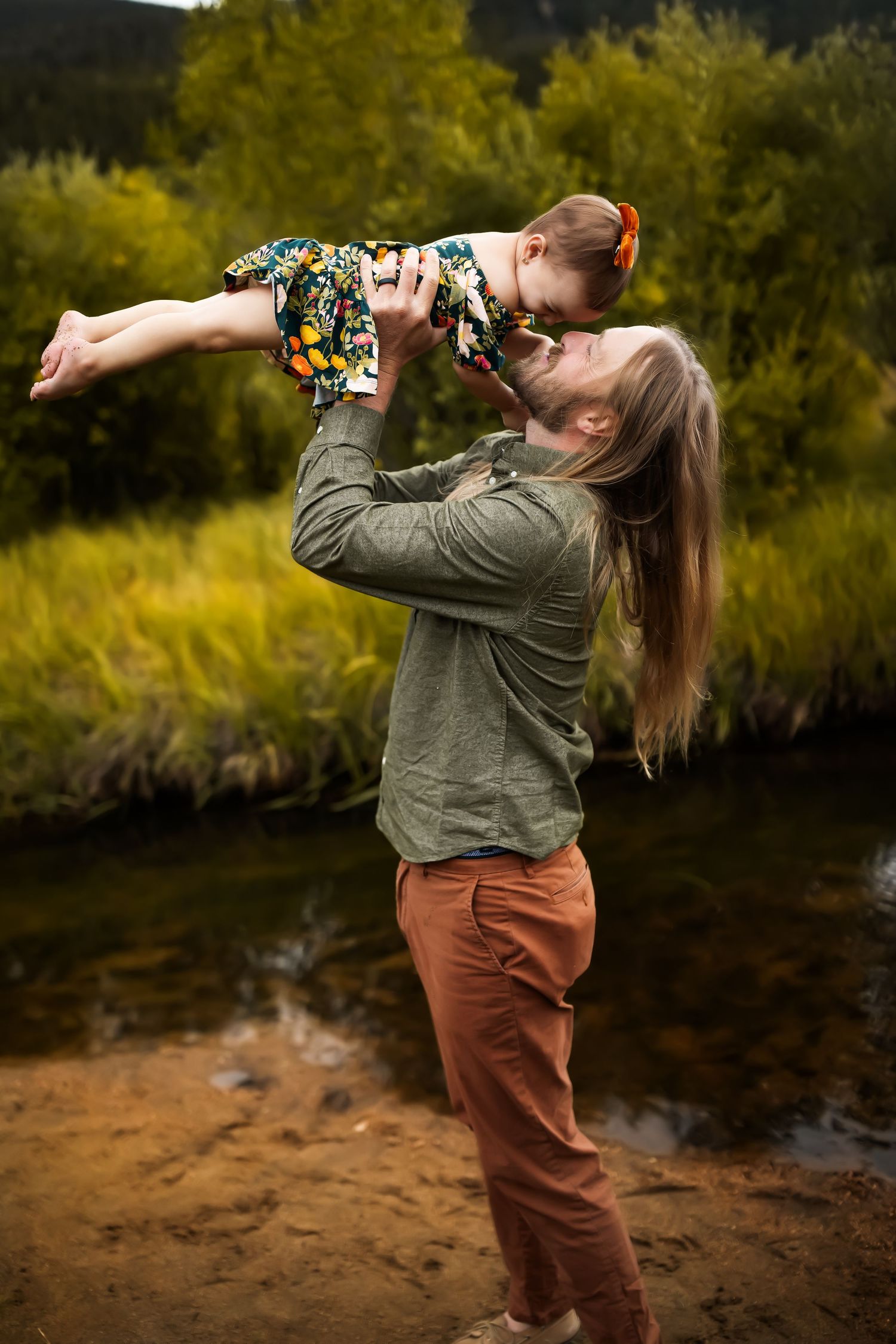 Parent lifting child wearing floral dress by riverside in autumn setting.