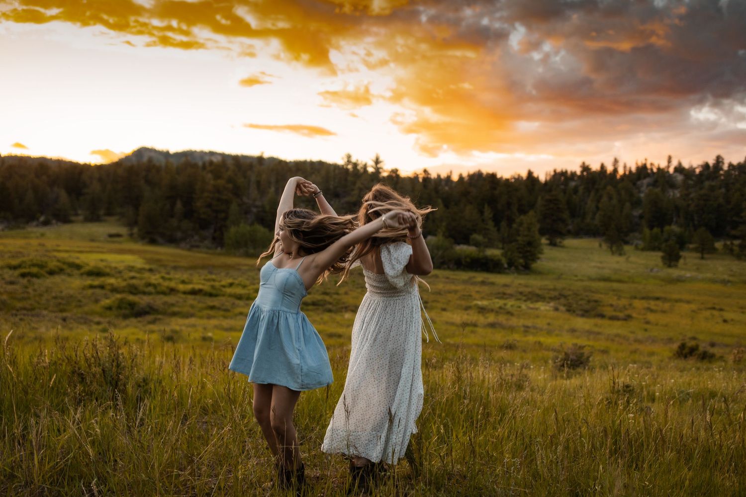 Two people in flowing dresses dancing together in a meadow at golden hour.