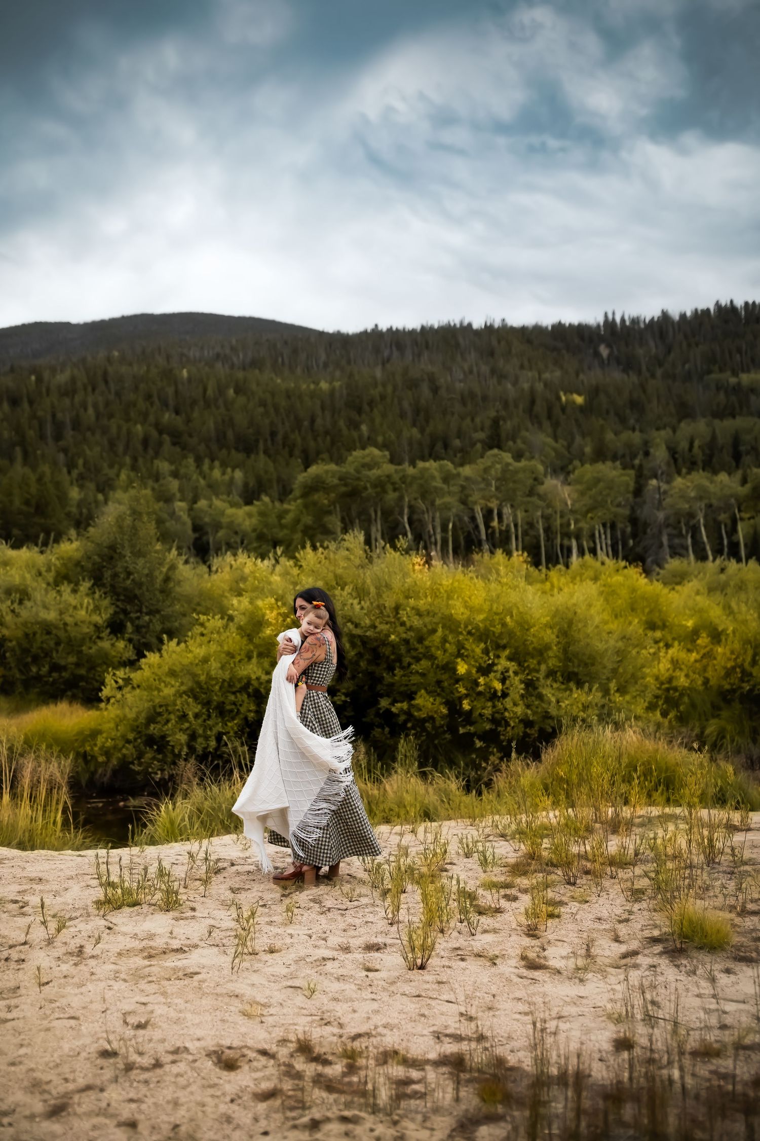 A couple embraces on a sandy beach area with mountains and forest in the background under moody storm clouds.