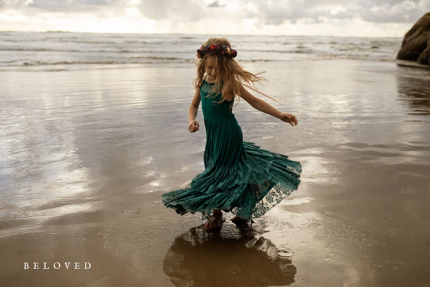 Flowing dress catches wind as figure twirls on moody beach at sunset.
