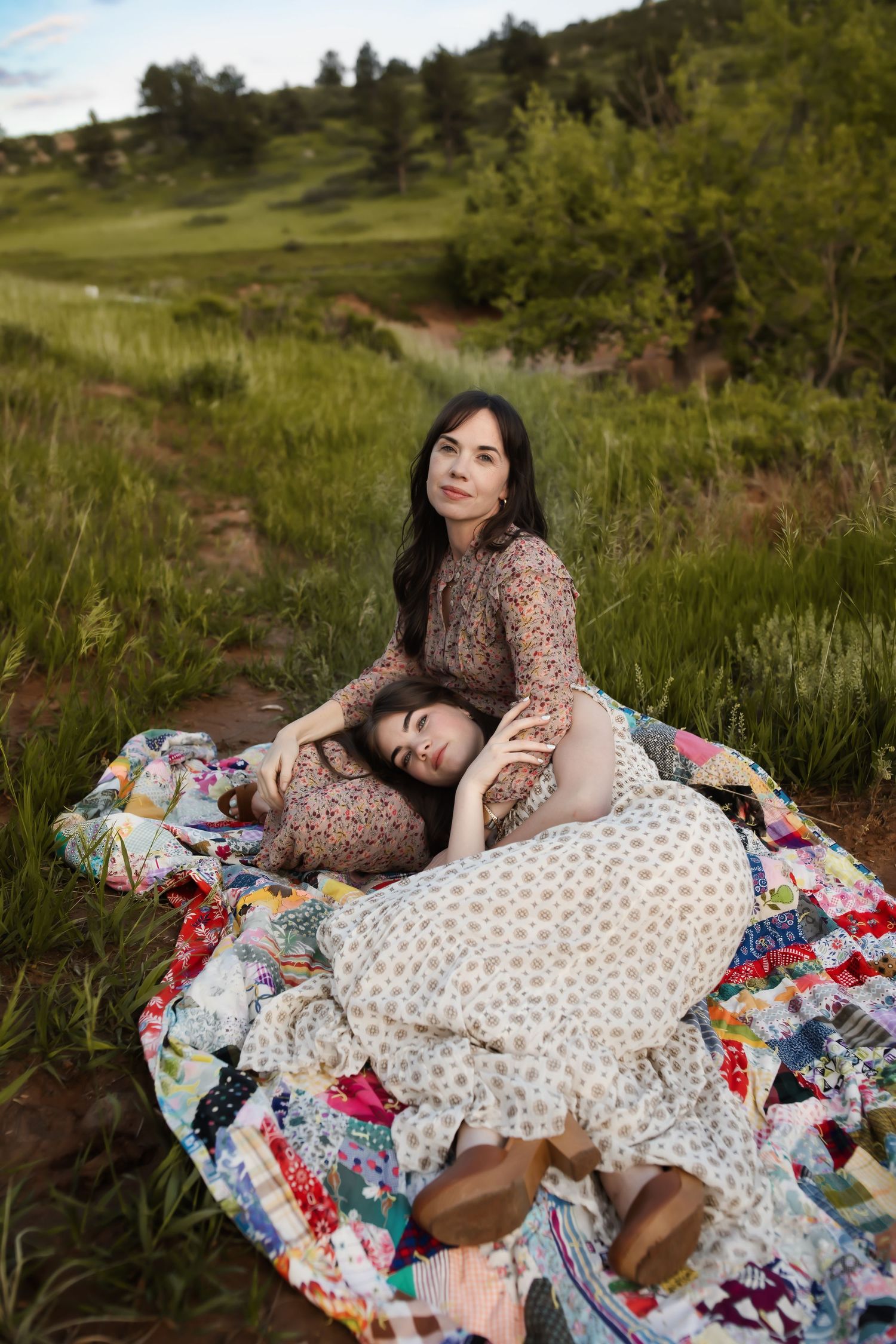 Two people resting on a colorful patchwork quilt in a grassy meadow setting.