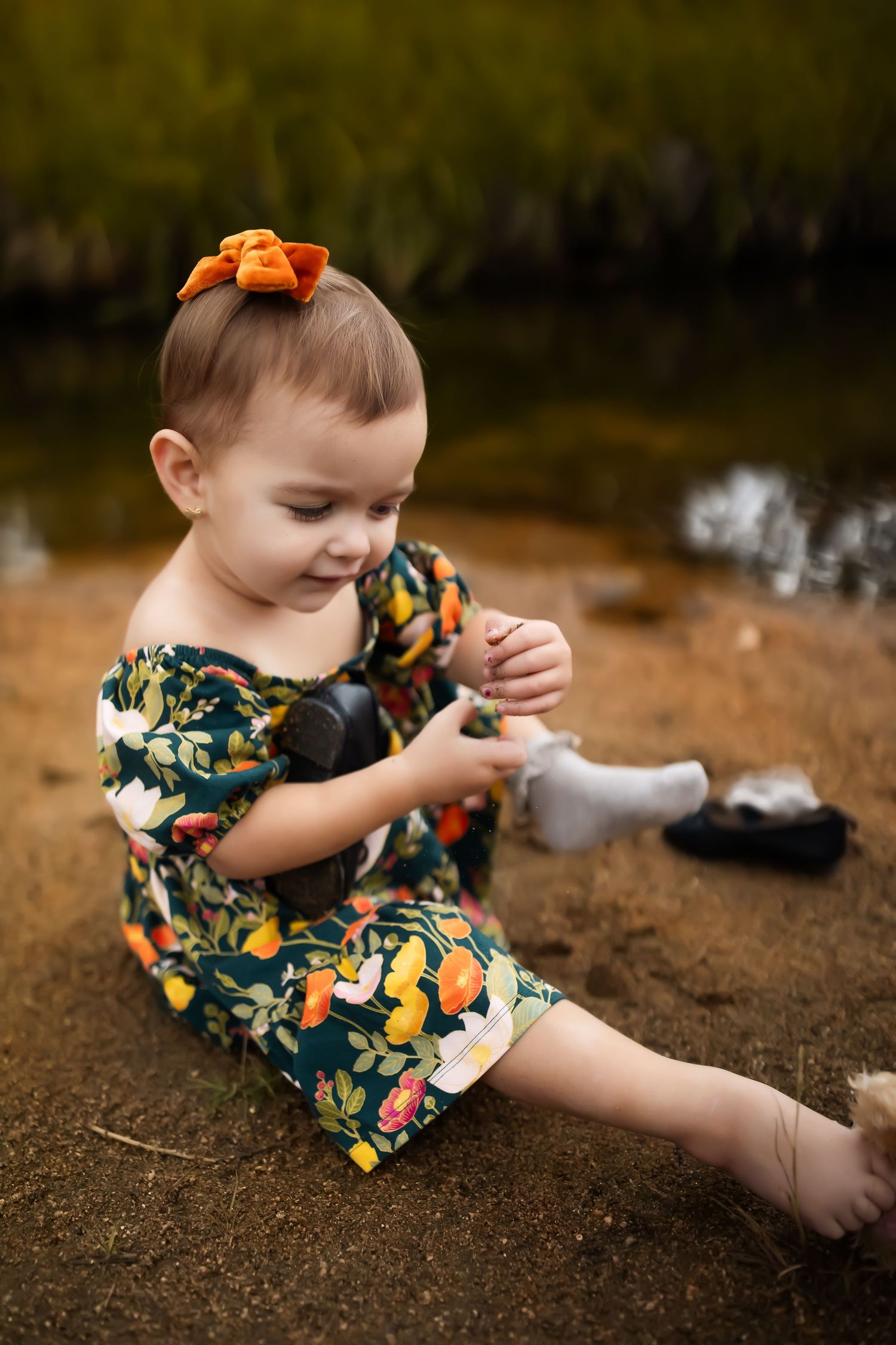 A toddler in a floral dress plays in the dirt near a river bank on an autumn day.