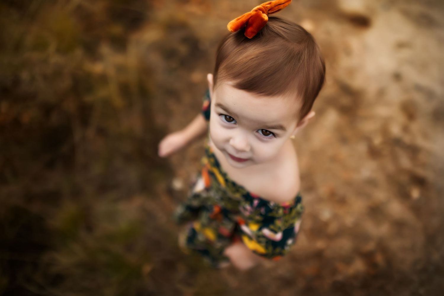 Baby in dark floral dress with orange hair bow smiling at camera against natural outdoor background.
