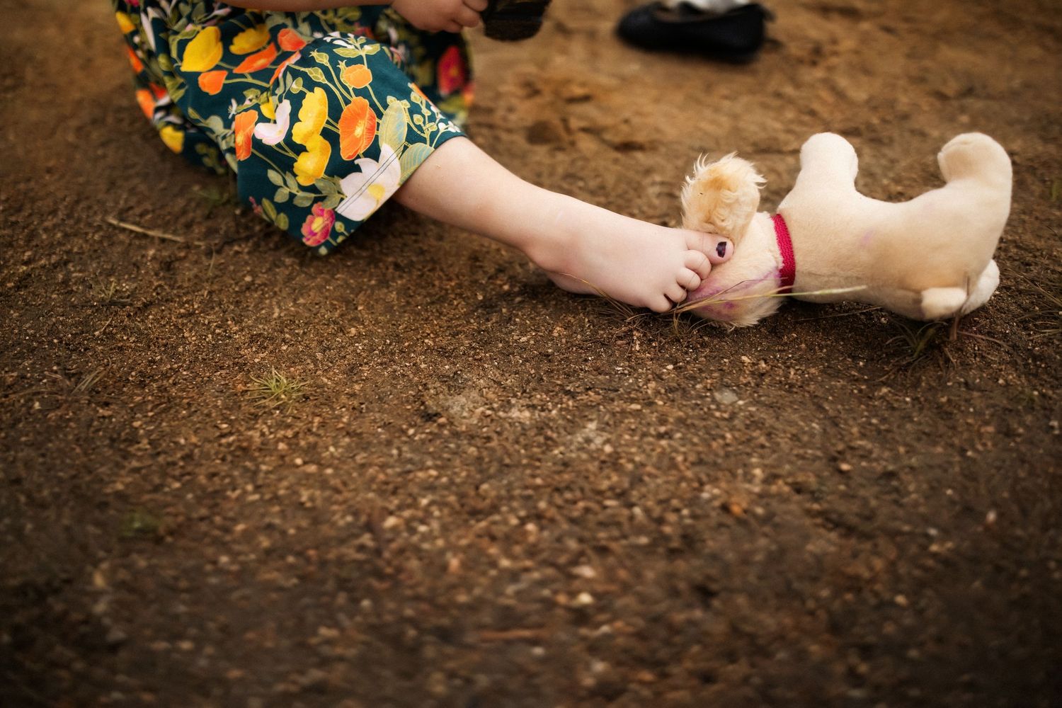 A small plush dog toy lies next to someone wearing a floral dress on brown dirt ground.